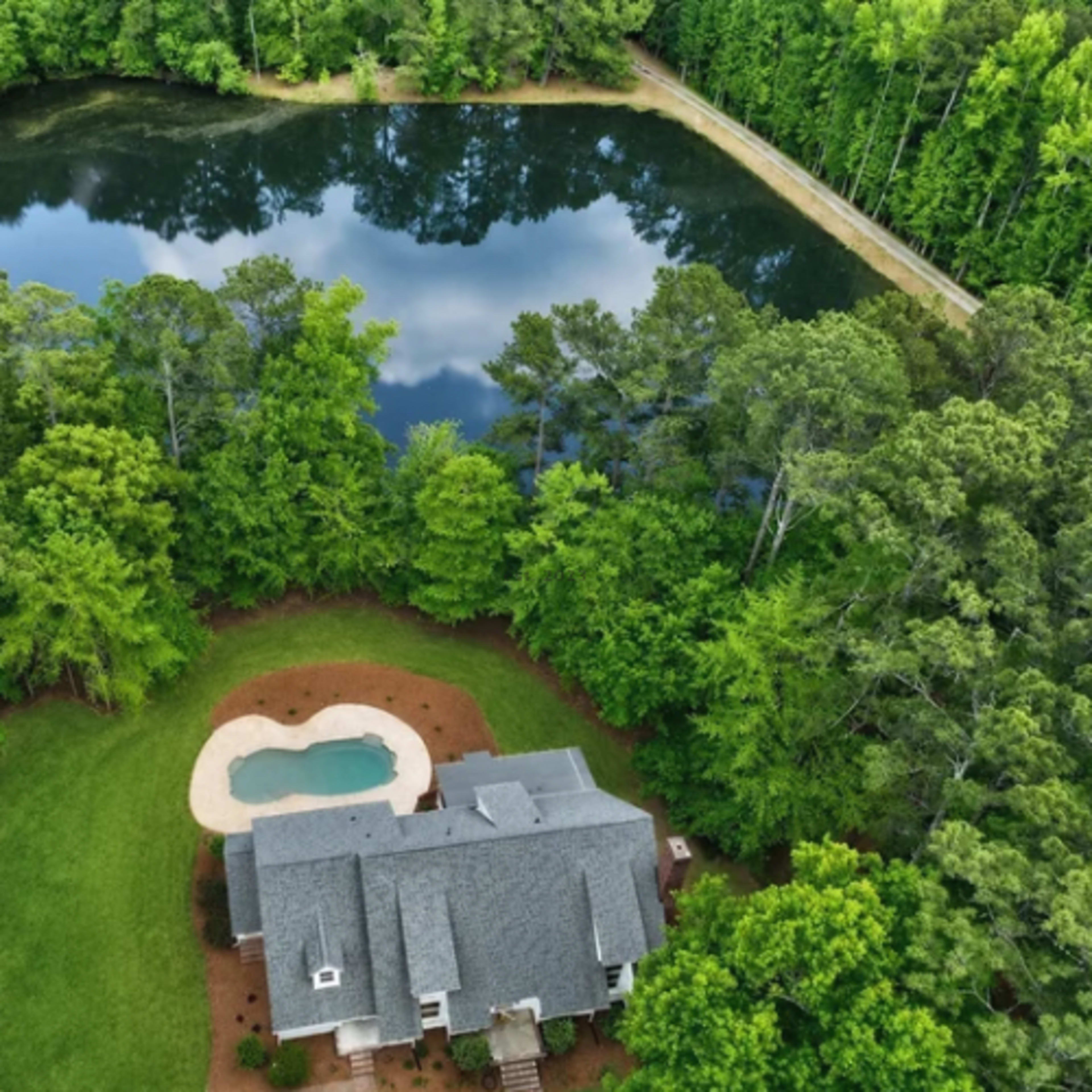 An aerial view shows a house with a swimming pool surrounded by trees and a pond reflecting the sky.