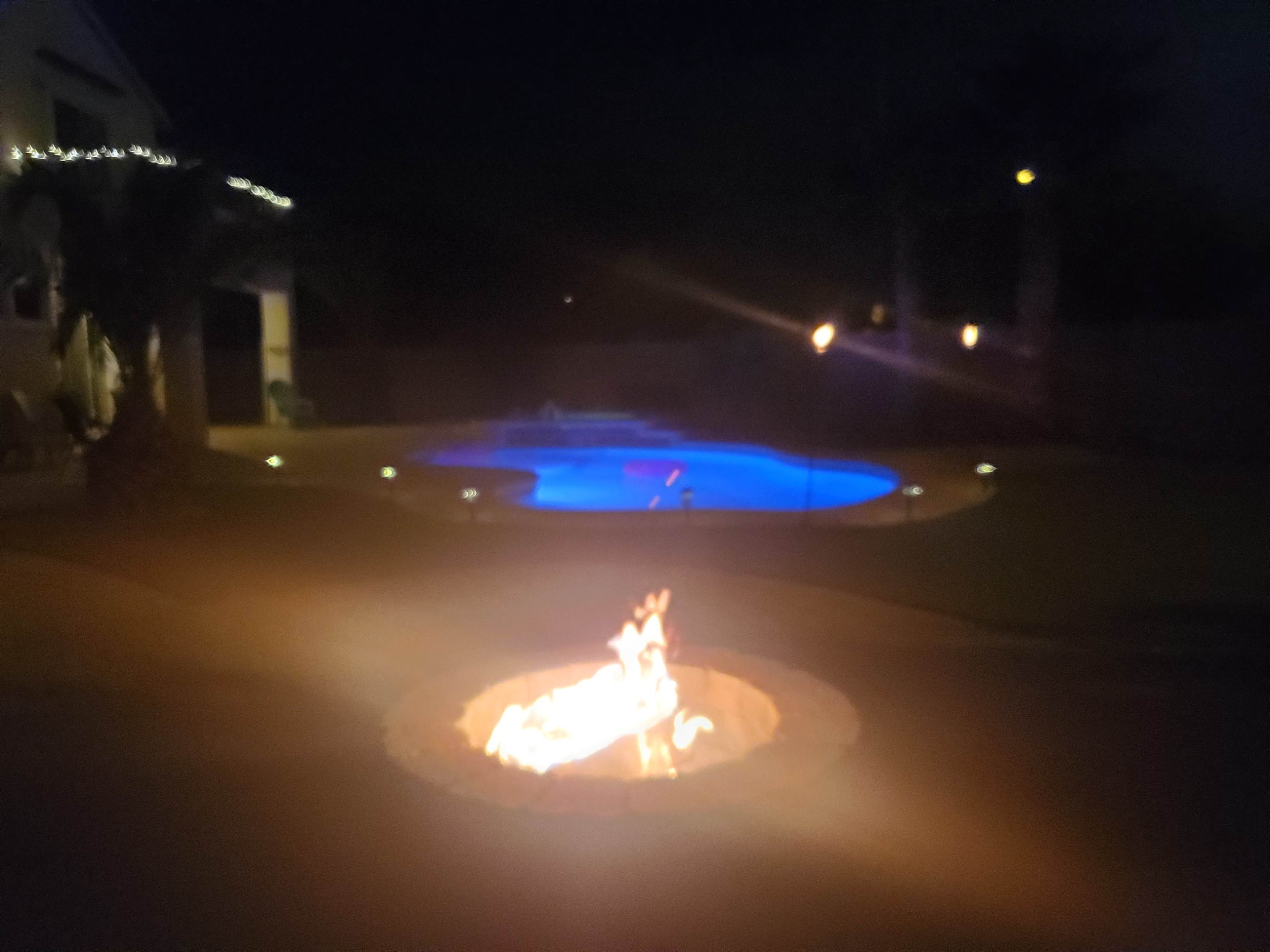 A fire pit with flames is in the foreground, while a lit swimming pool is visible in the background under a night sky.