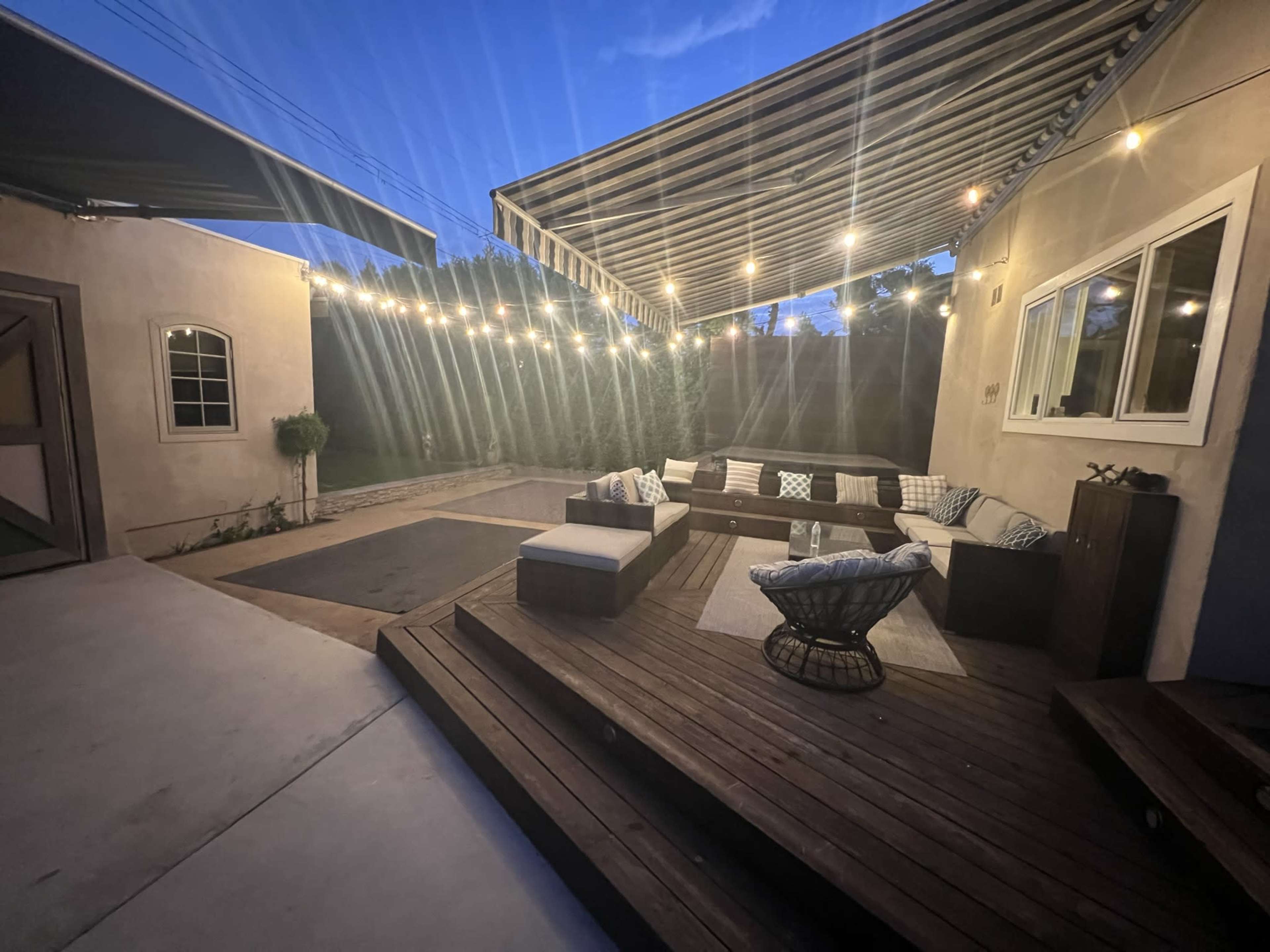 The image shows a patio area with wooden decking, outdoor seating, and string lights under a canopy at dusk.