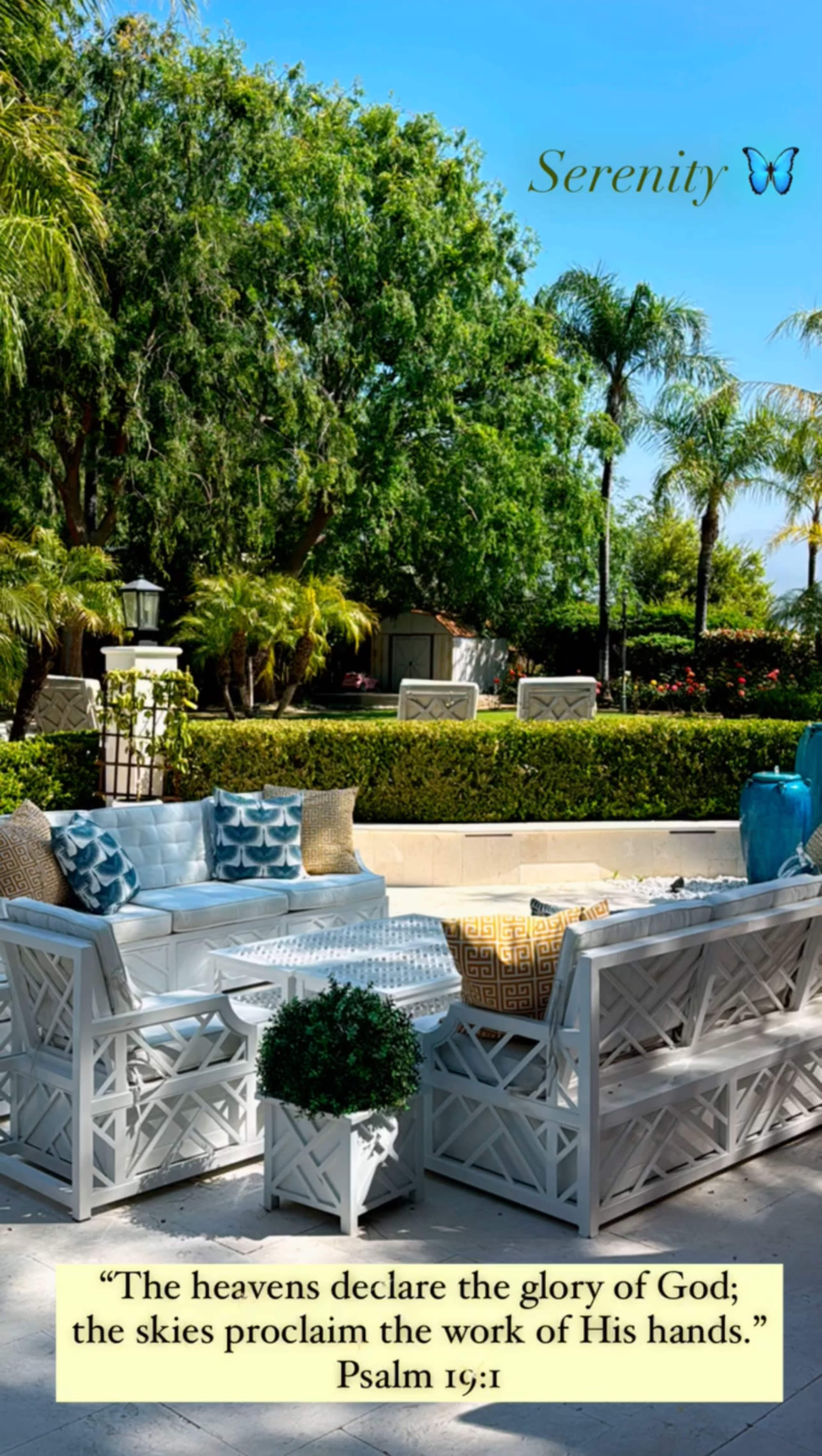 A well-decorated outdoor seating area features a white wicker sofa, colorful cushions, and lush greenery under a clear blue sky.