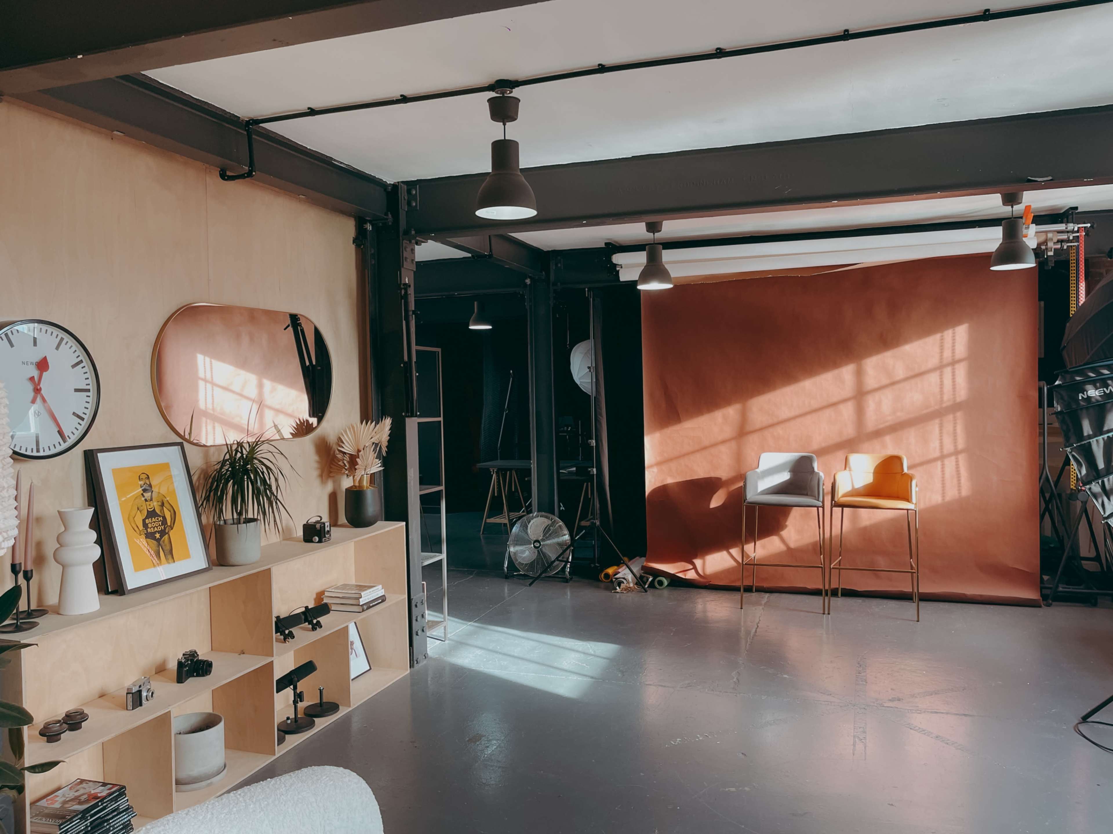 The image shows a photography studio with a wooden wall, a large mirror, a shelf with decorative items and cameras, and a backdrop featuring orange fabric.