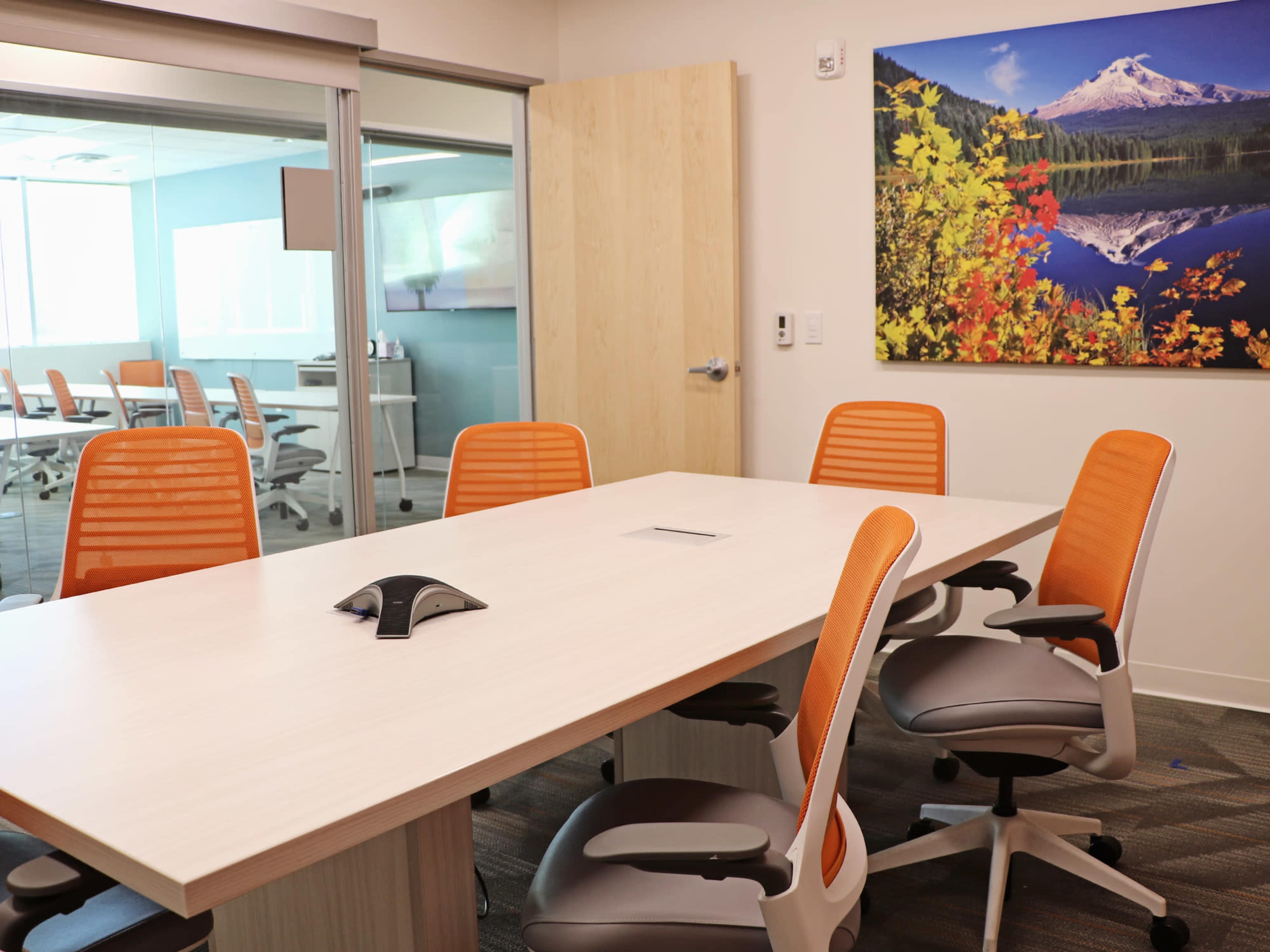 A modern conference room with a rectangular table, four orange office chairs, and a large wall-mounted nature landscape artwork.