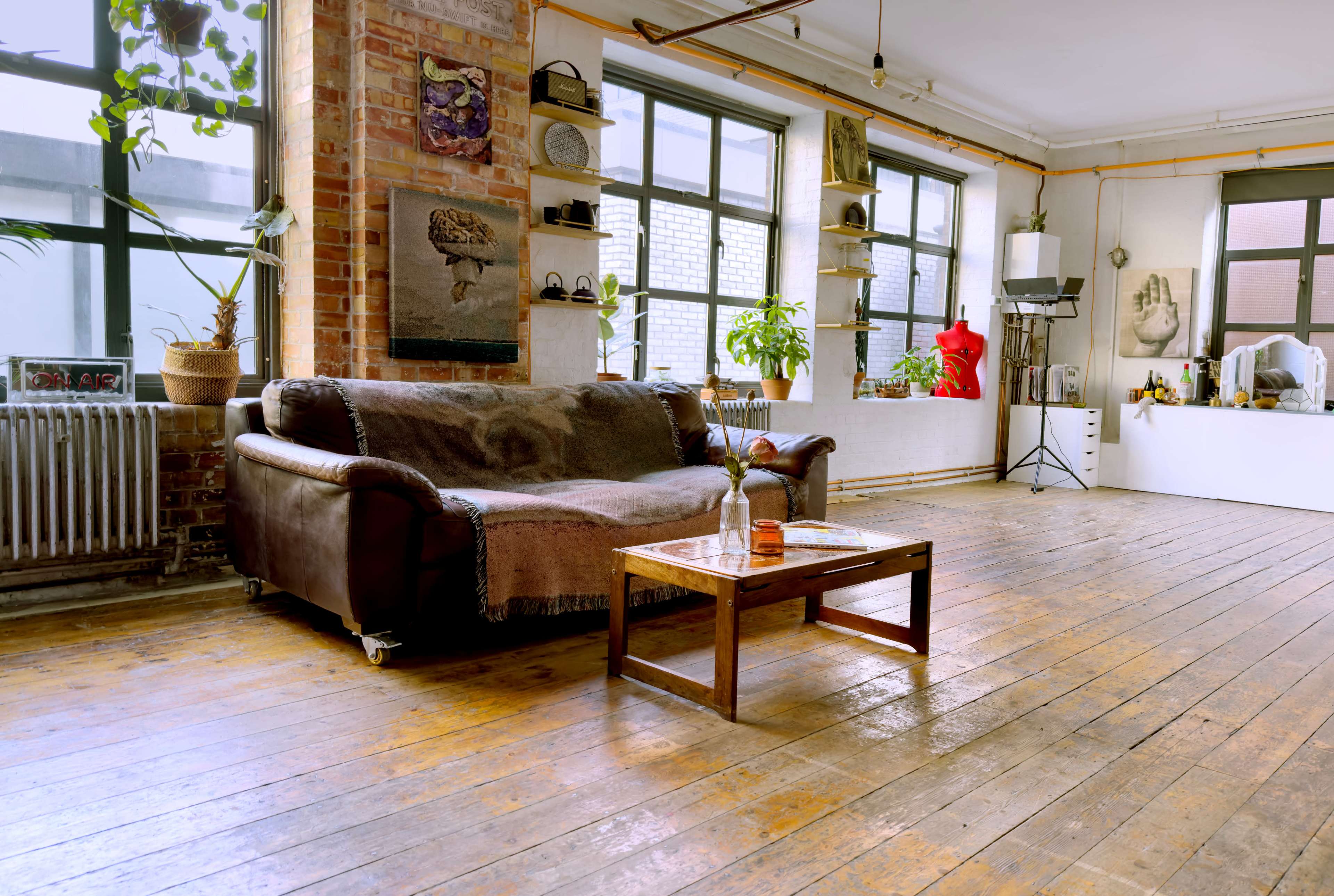 A spacious, sunlit room featuring a brown leather sofa, a wooden coffee table, and large windows with plants and decorative items placed throughout.