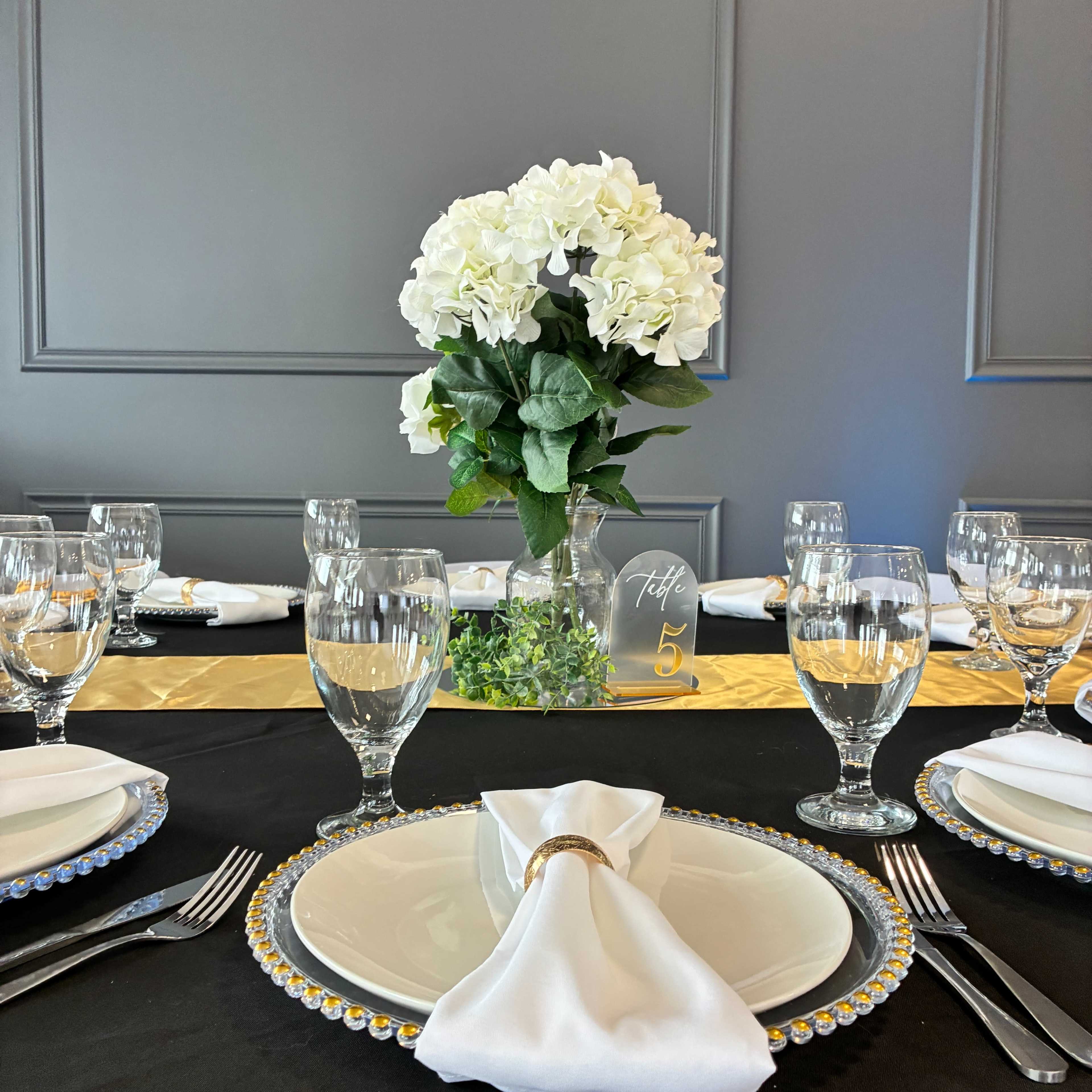 A dining table is elegantly set with white plates, crystal-clear glasses, and a floral centerpiece, surrounded by gold and black decor elements.