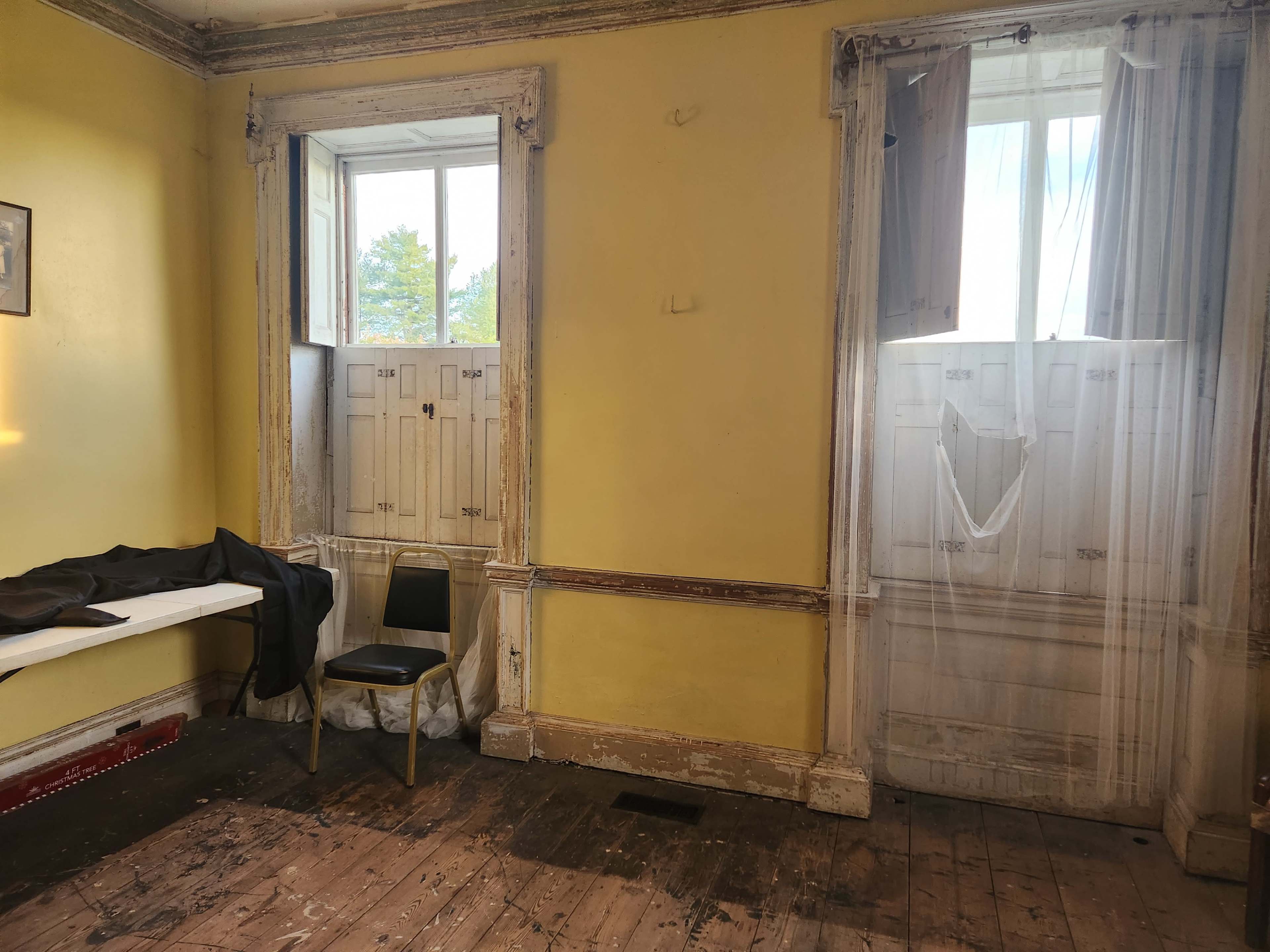A room with yellow walls, two windows with wooden shutters, a black garment draped over a white table, and a single black chair on a wooden floor.