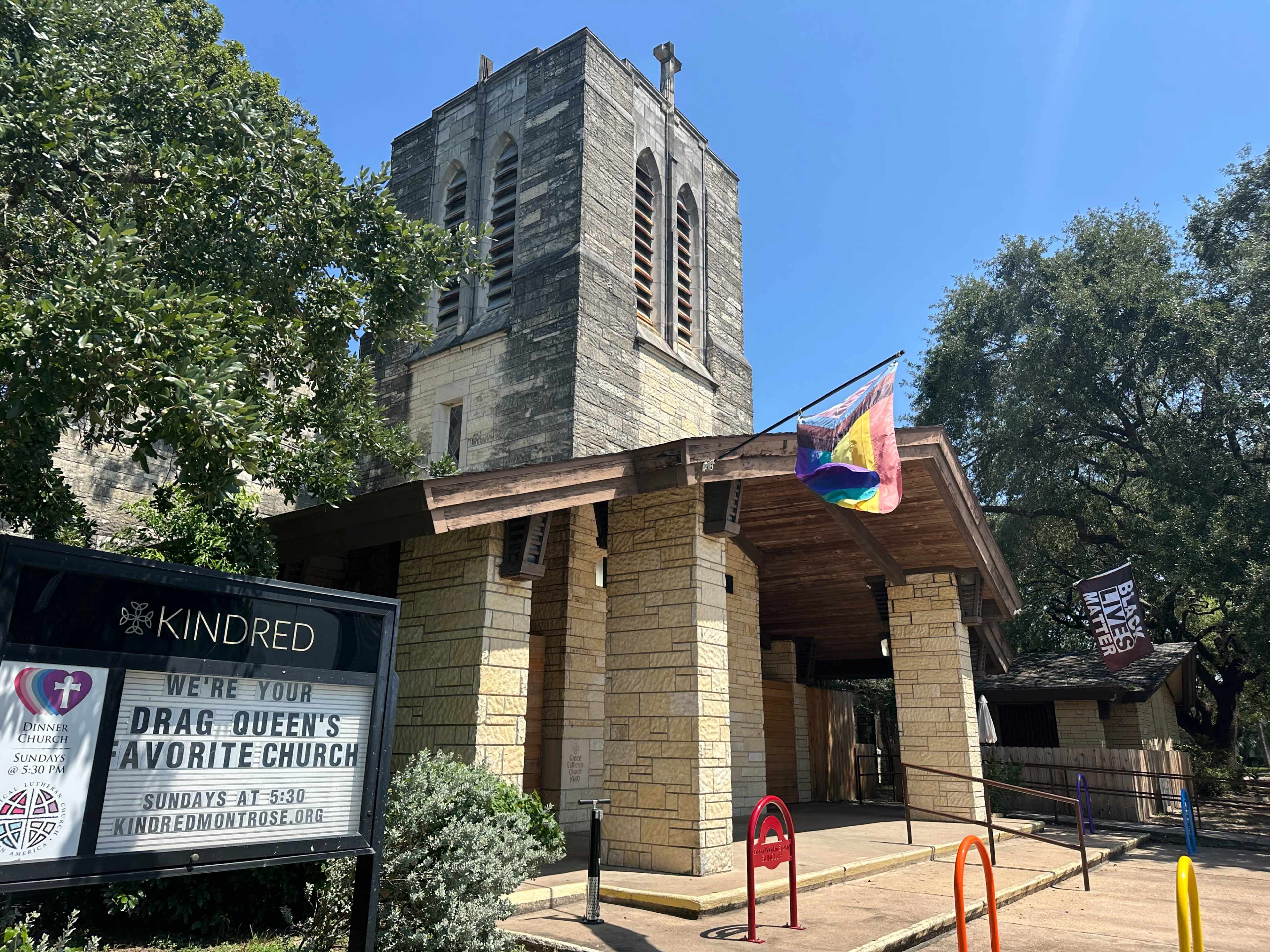 The image shows a church building with a stone façade, featuring a bell tower and a colorful pride flag hanging from the entrance.