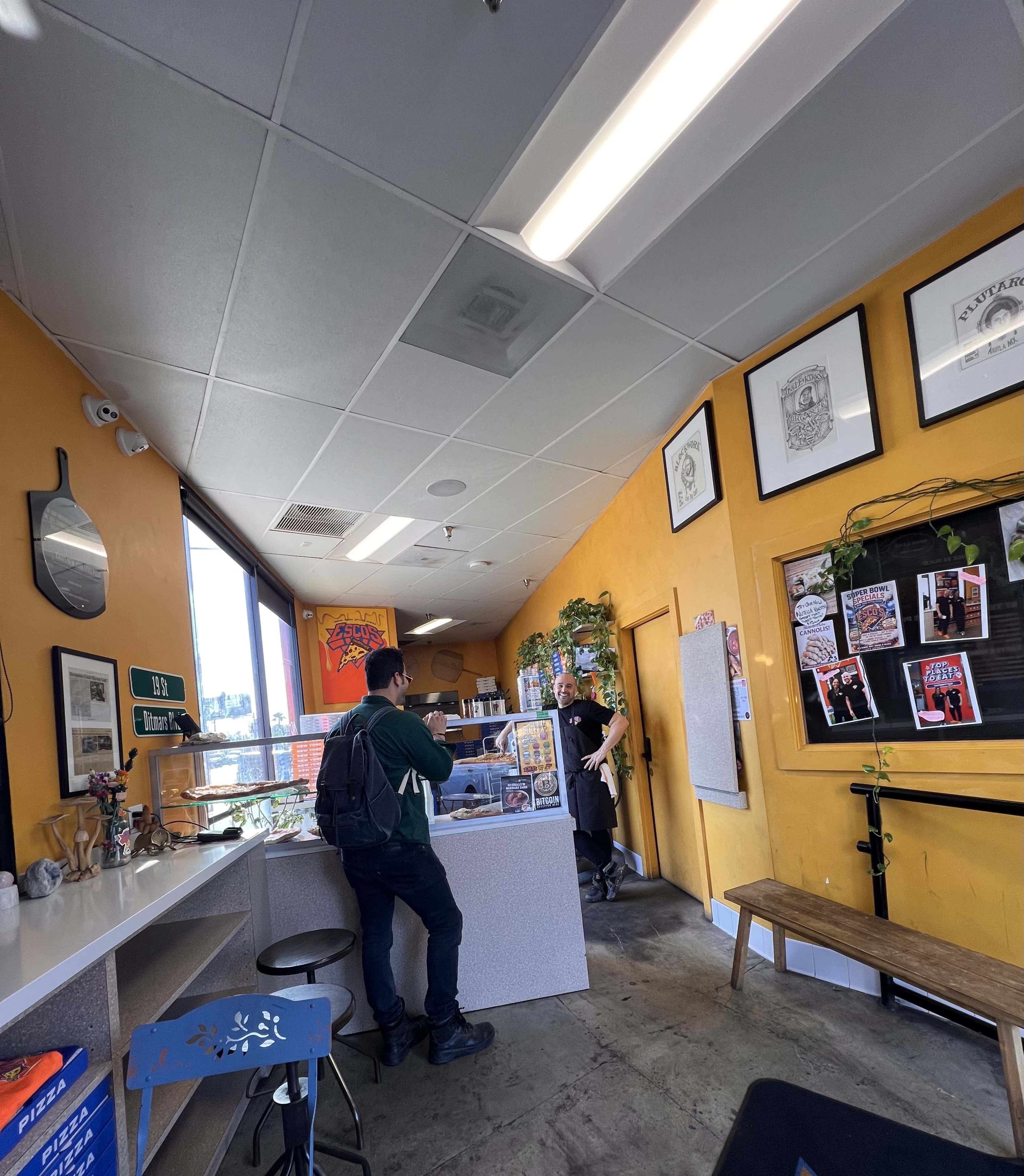 A person stands at a counter while another person, presumably a staff member, engages with them in a brightly colored café adorned with various wall decorations.