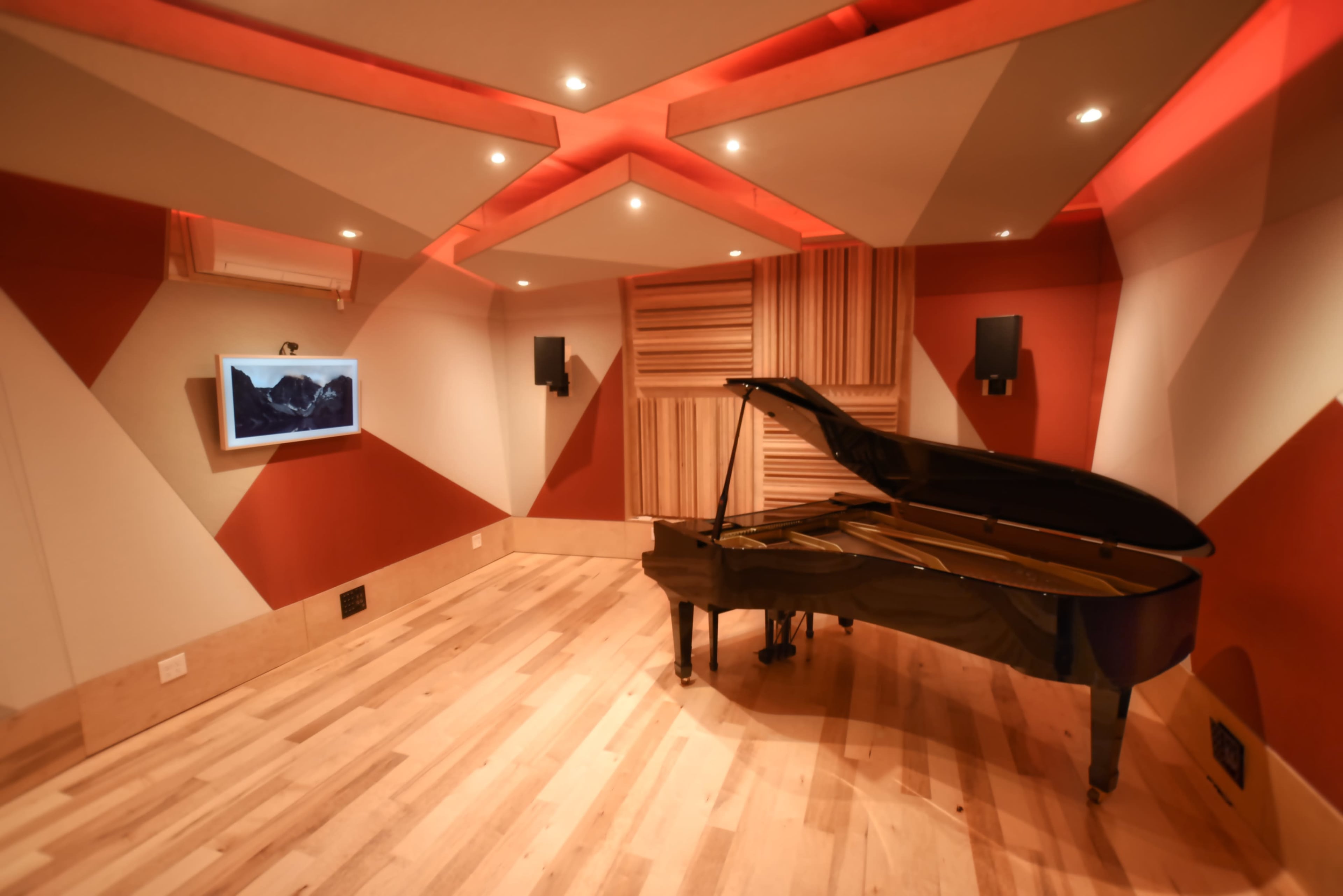 A black grand piano is positioned in a soundproof room with patterned red and beige walls, wooden flooring, and ambient lighting.
