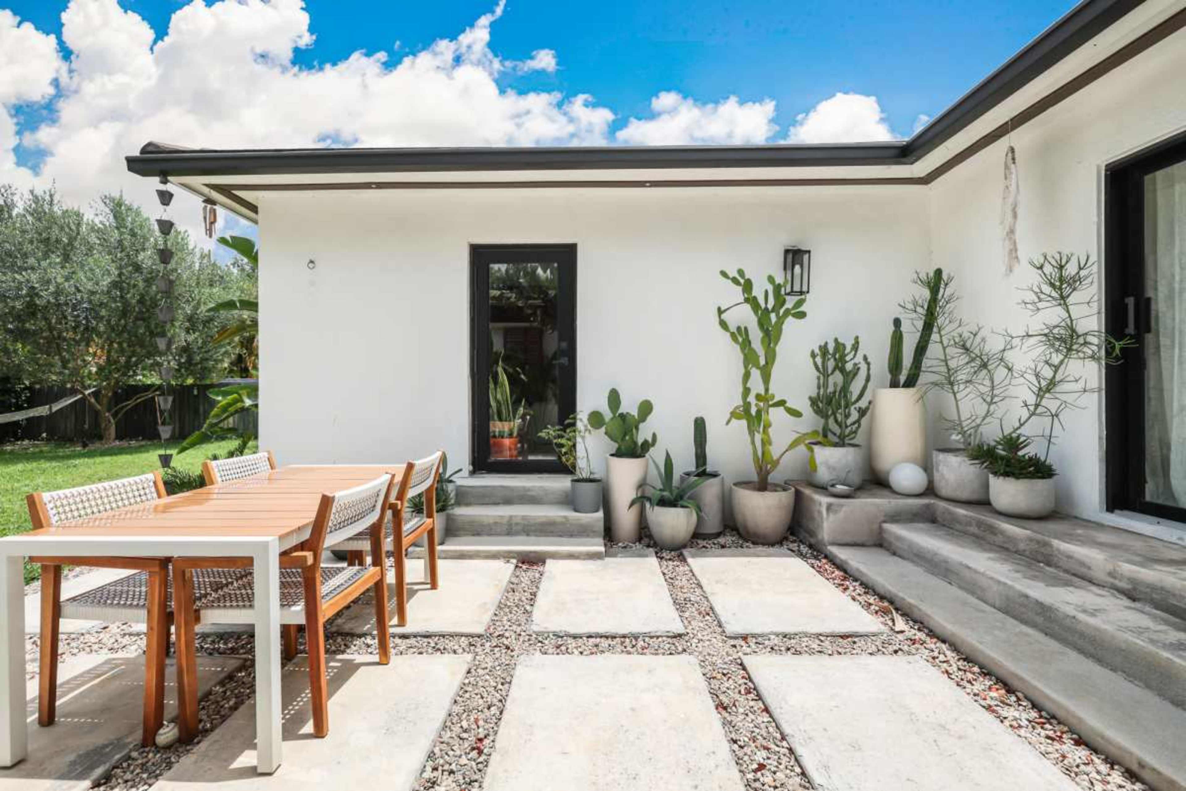A modern outdoor patio features a wooden dining table and chairs surrounded by various potted plants and cacti against a white wall.