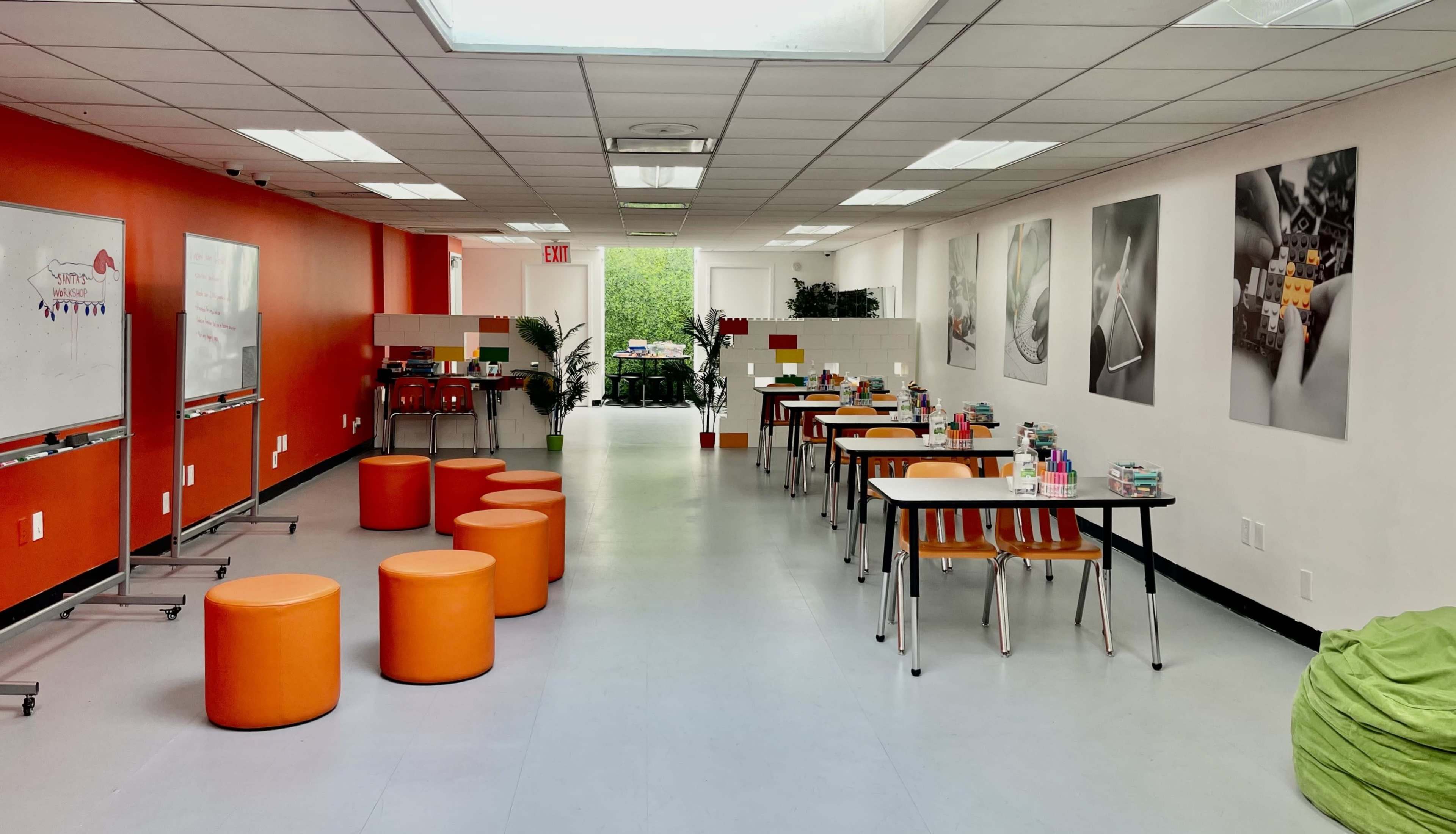 A brightly lit room with orange accents, featuring tables and stools arranged in two rows along the walls, and plants near the entrance.
