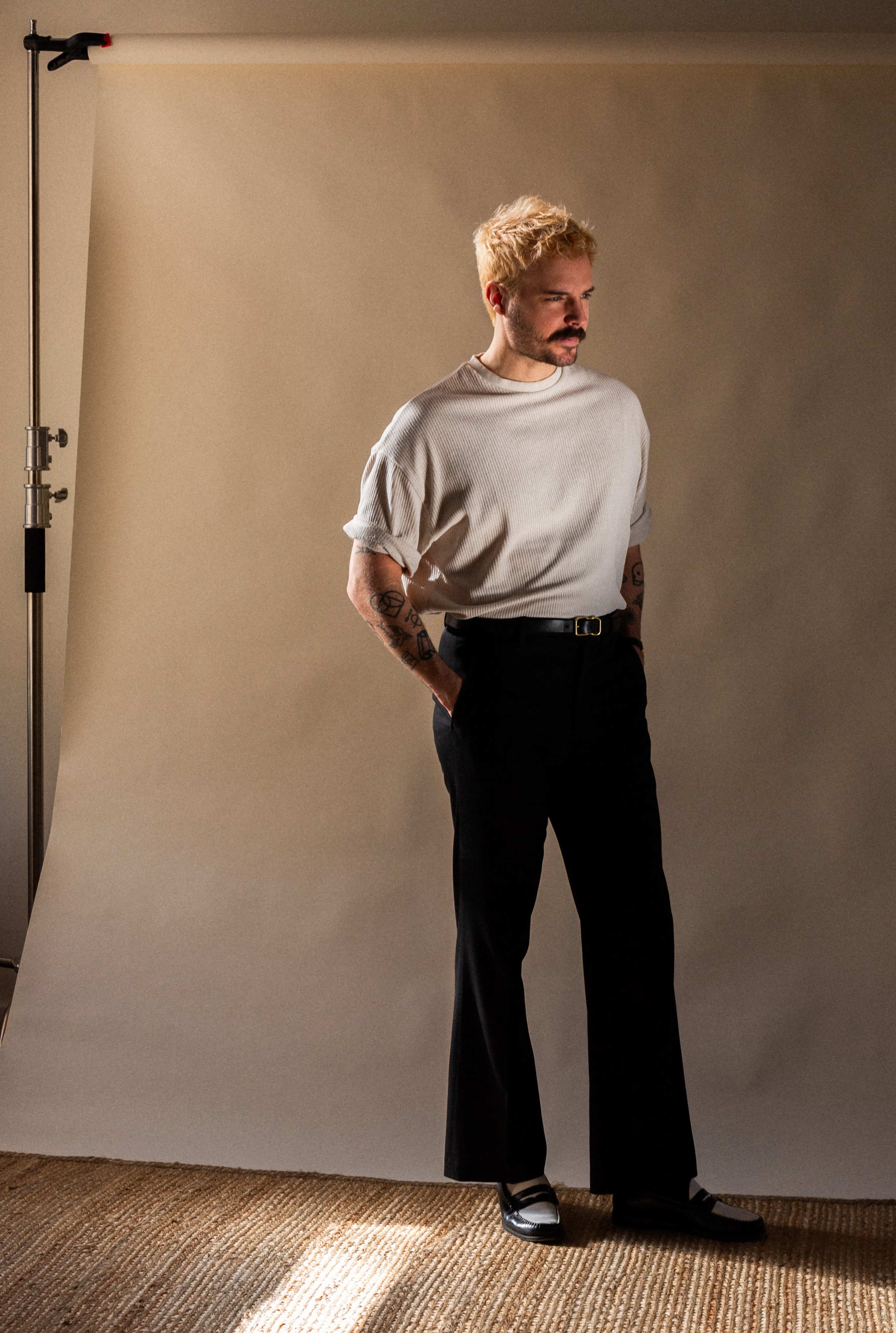 A man stands in front of a light gray backdrop, wearing a white shirt and black trousers with a belt, while his hands rest in his pockets.