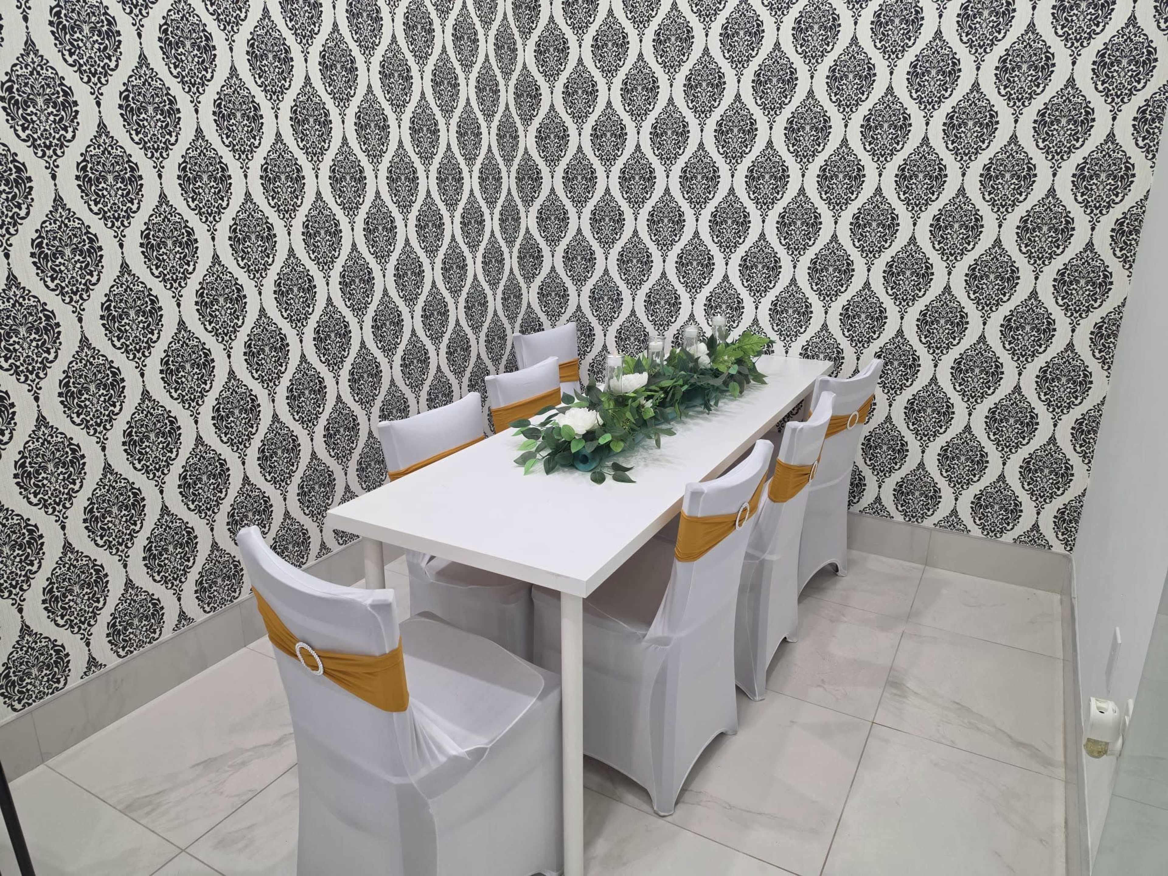 A white table is surrounded by eight chairs covered in white and gold sashes, positioned in a room with a patterned black and white wallpaper.
