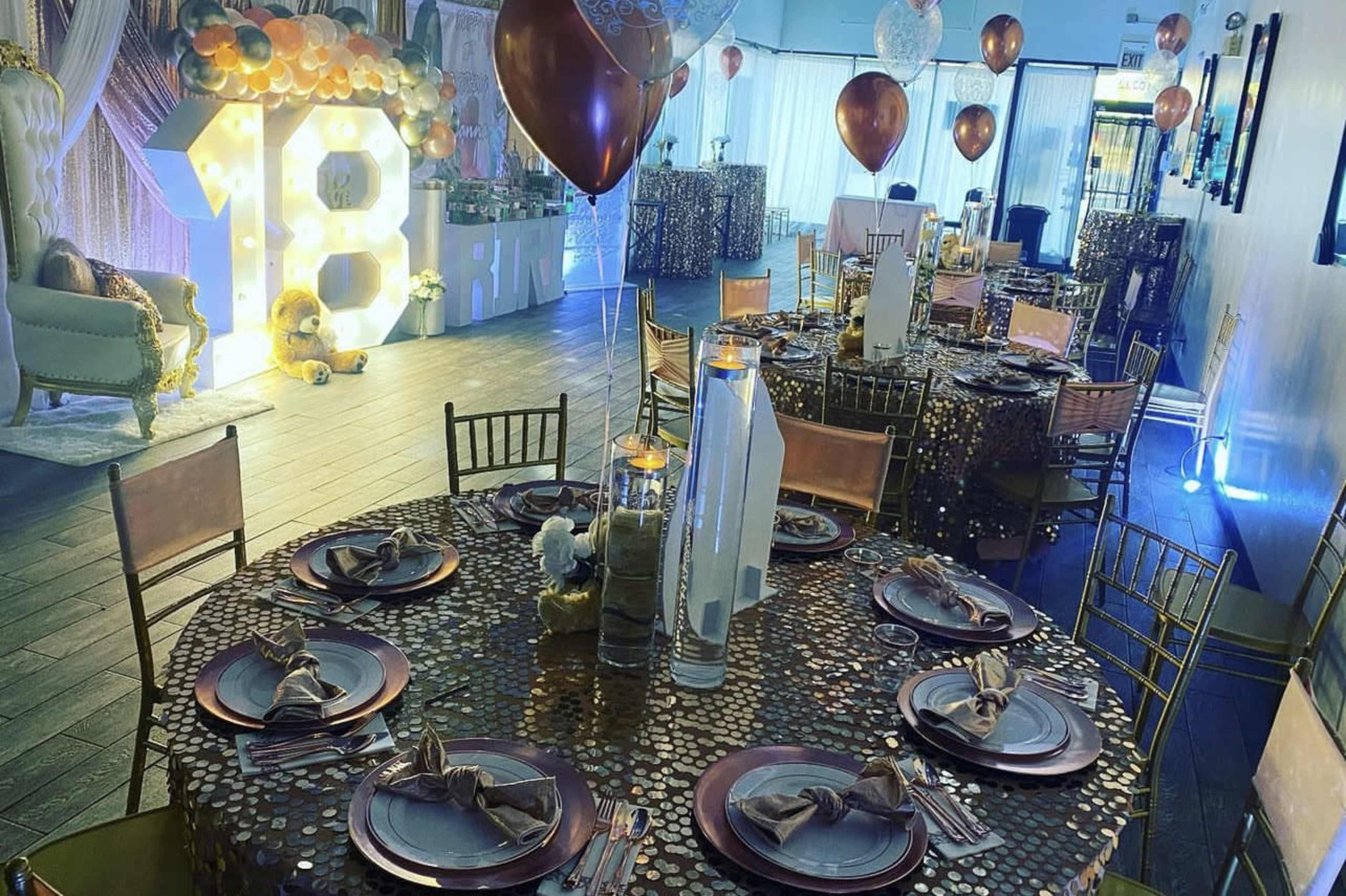 A decorated banquet hall with tables set for a celebration, featuring golden tablecloths, centerpieces, and balloons.