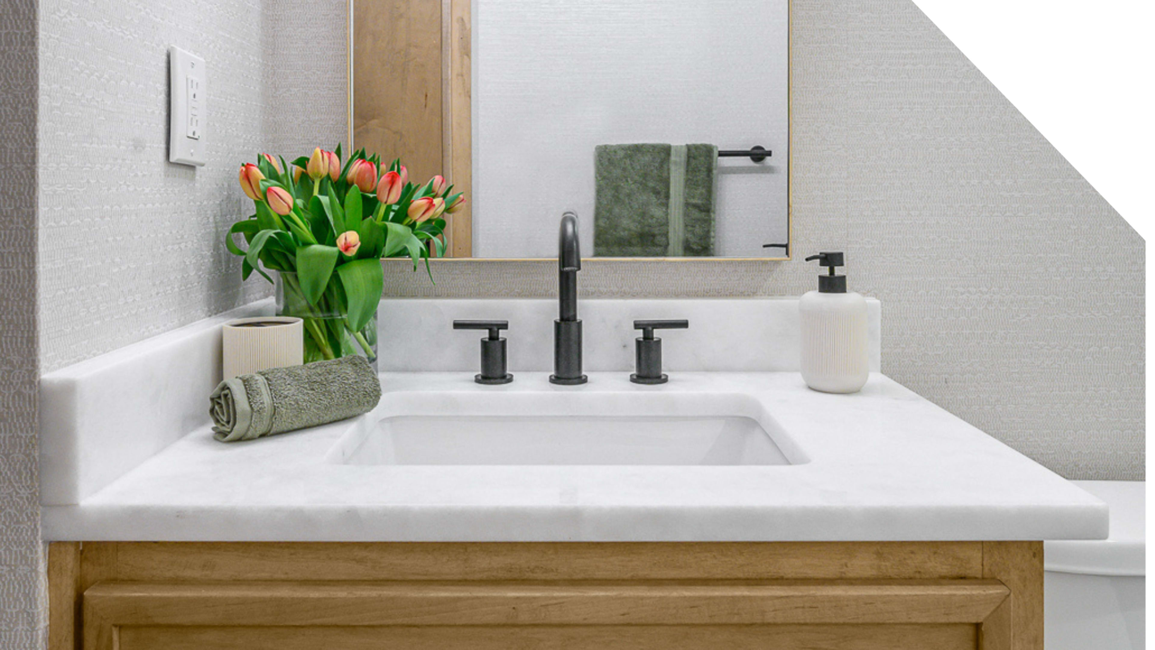 A bathroom countertop features a vessel sink, faucet set, a small vase of tulips, a rolled towel, a soap dispenser, and a hand towel near a framed mirror.
