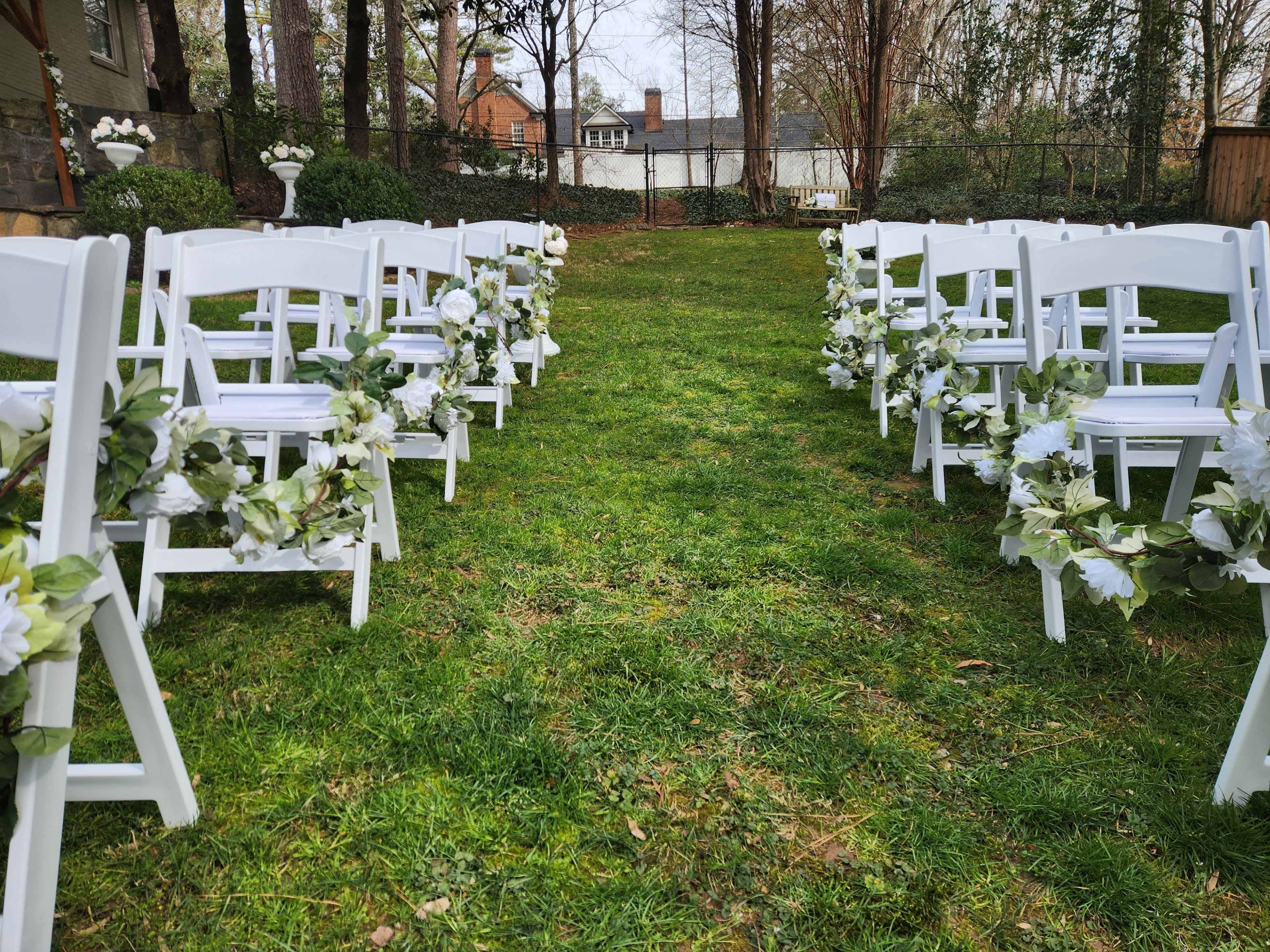 A grassy outdoor area set up for a wedding ceremony, with white folding chairs lined up along a path decorated with greenery and flowers.