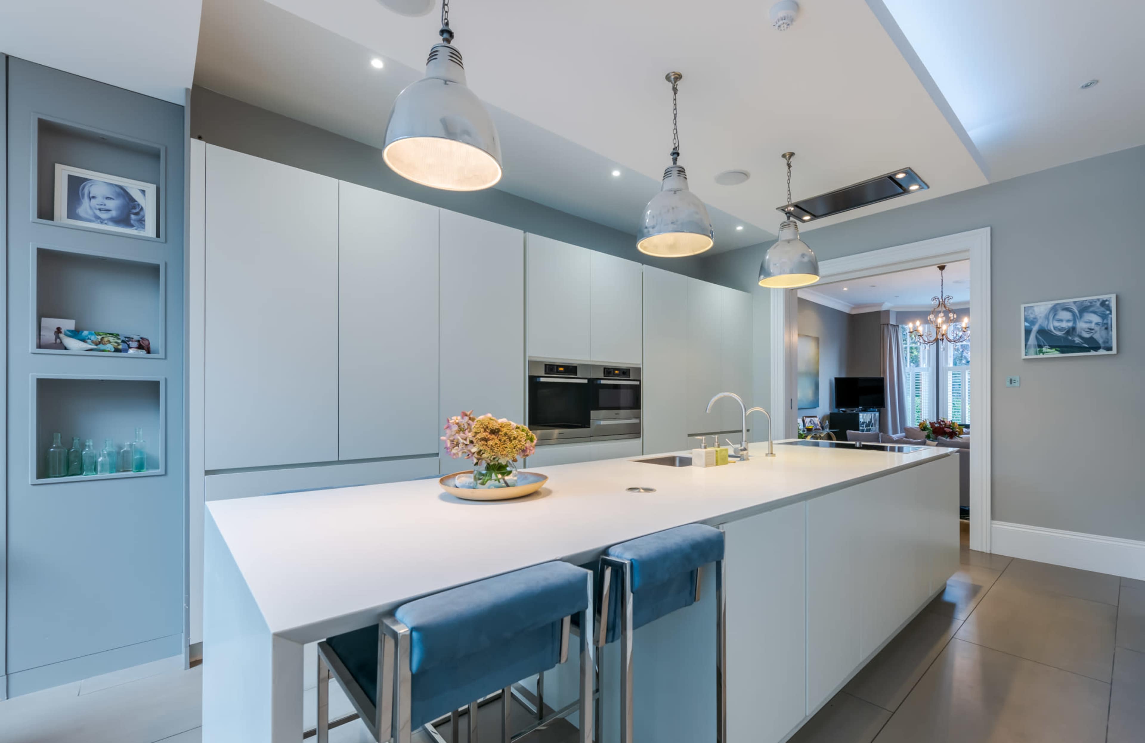 A modern kitchen features a central island with two stools, built-in appliances, and pendant lighting.