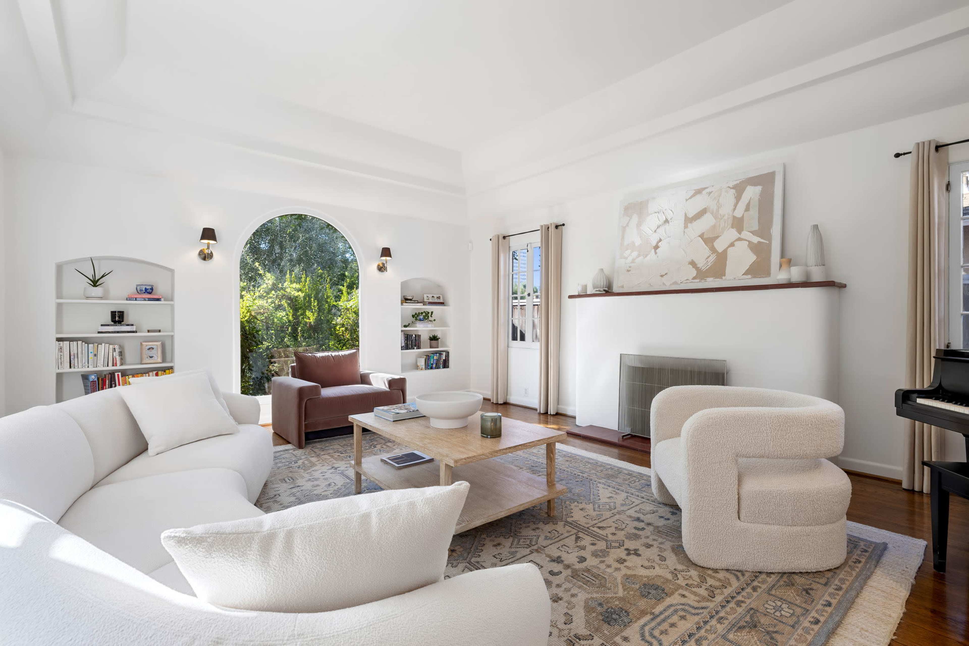 A modern living room features a white sectional couch, two armchairs, a wooden coffee table, and large windows with views of greenery.