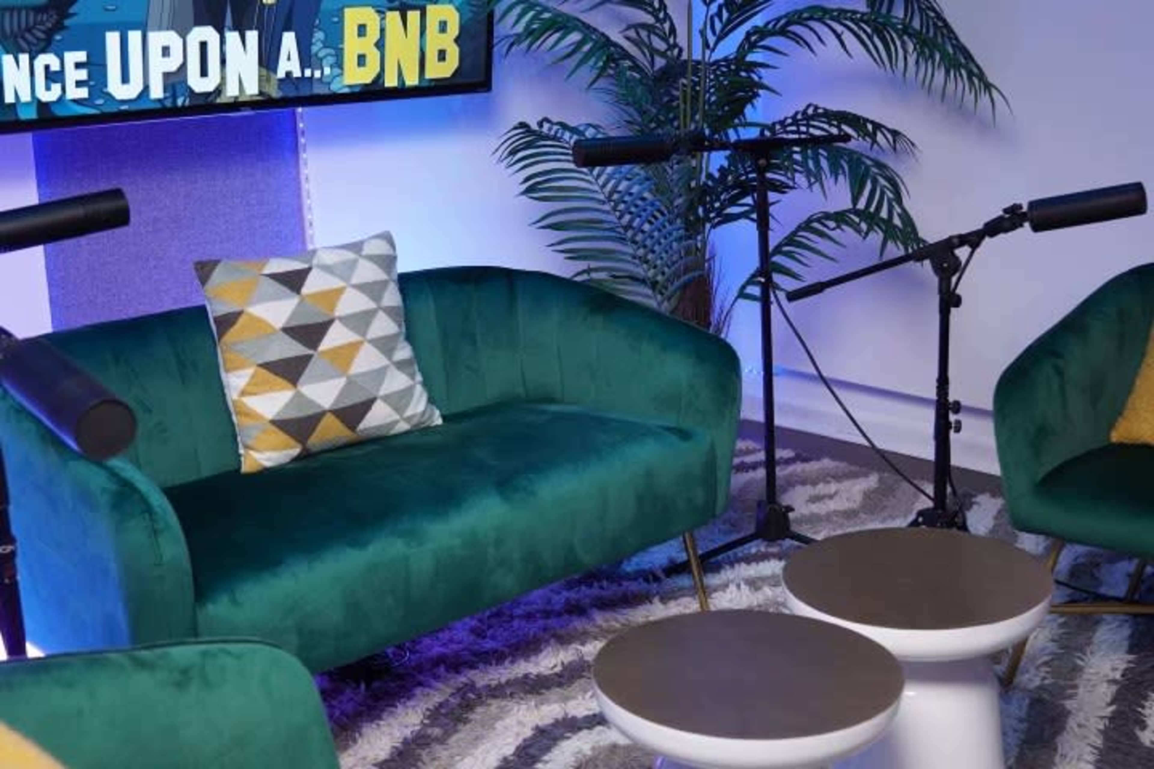 A green velvet couch with a geometric-patterned pillow, accompanied by two small white tables and microphones on either side, set against a backdrop with blue lighting and a plant.