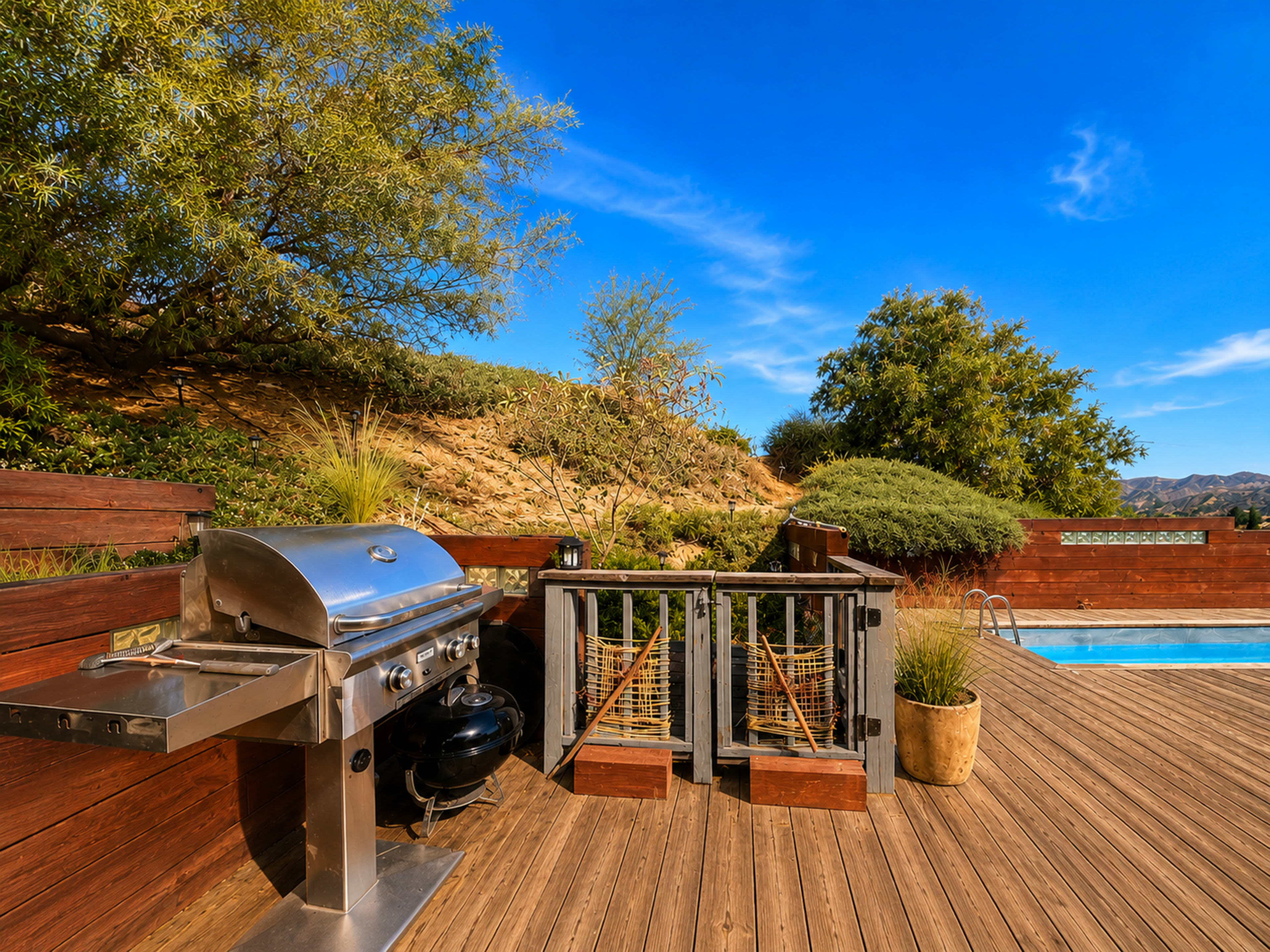 The image shows a wooden deck featuring a stainless steel barbecue grill, a railing with decorative accents, and a swimming pool surrounded by landscaped greenery under a clear blue sky.