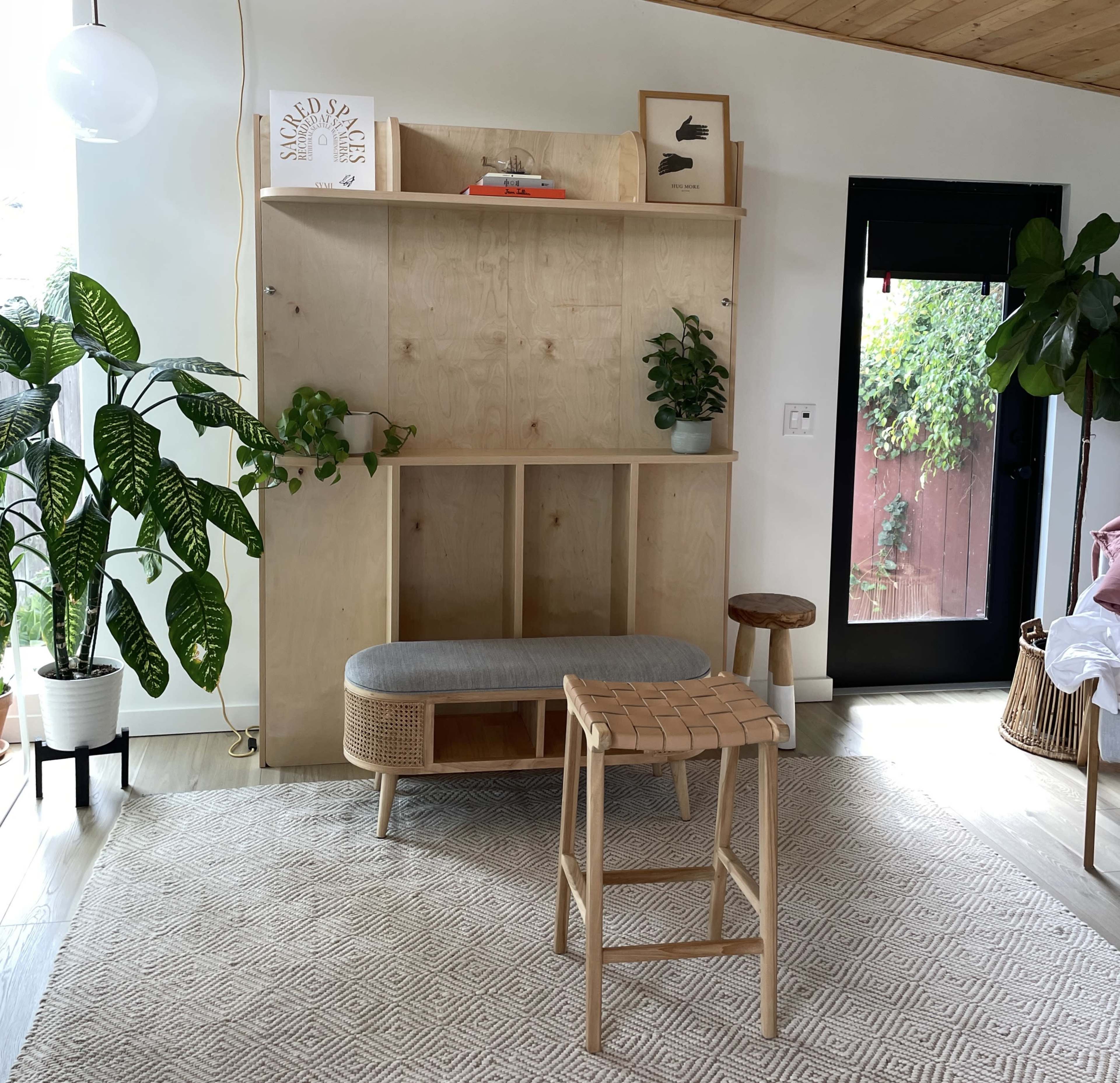 A modern interior space featuring a wooden storage unit with plants, a gray bench, and a woven stool on a patterned rug.