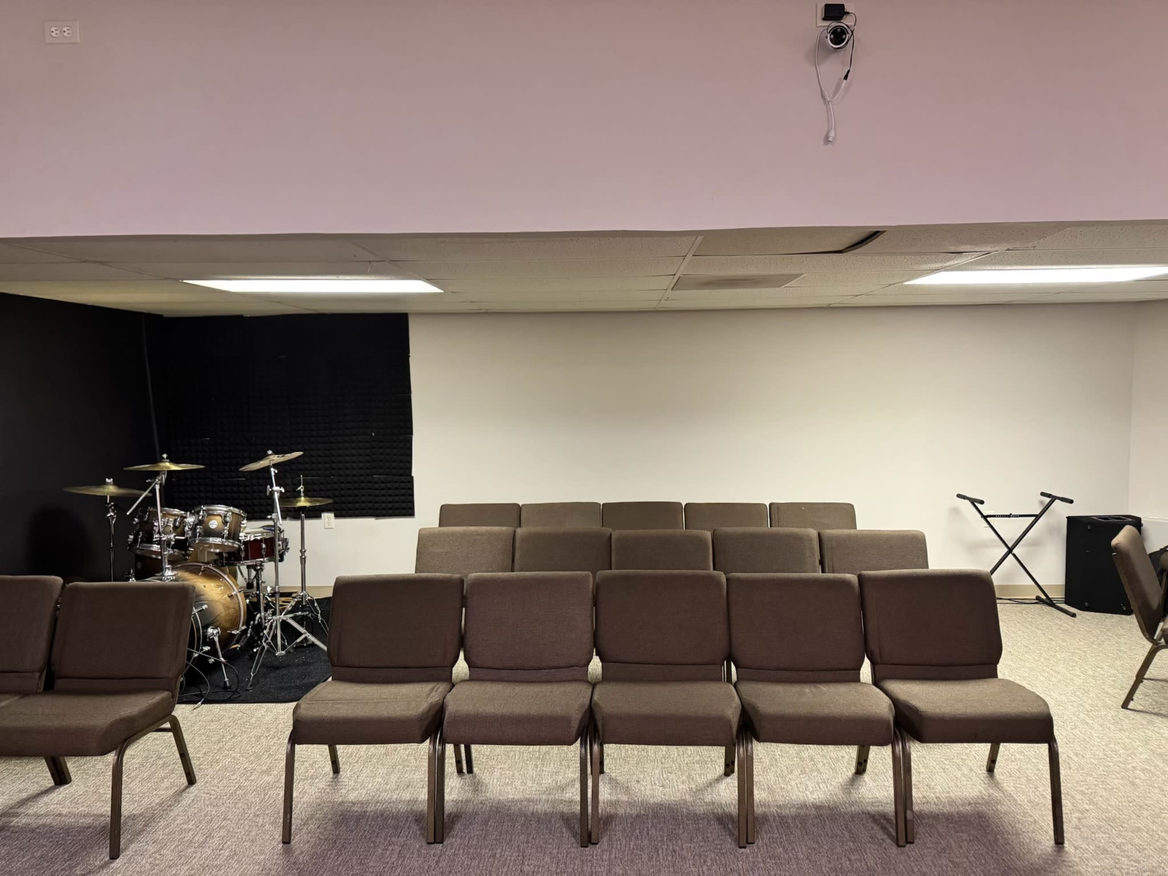 The image shows a well-lit room with rows of chairs facing a blank wall, a drum set in the corner, and a keyboard stand nearby.