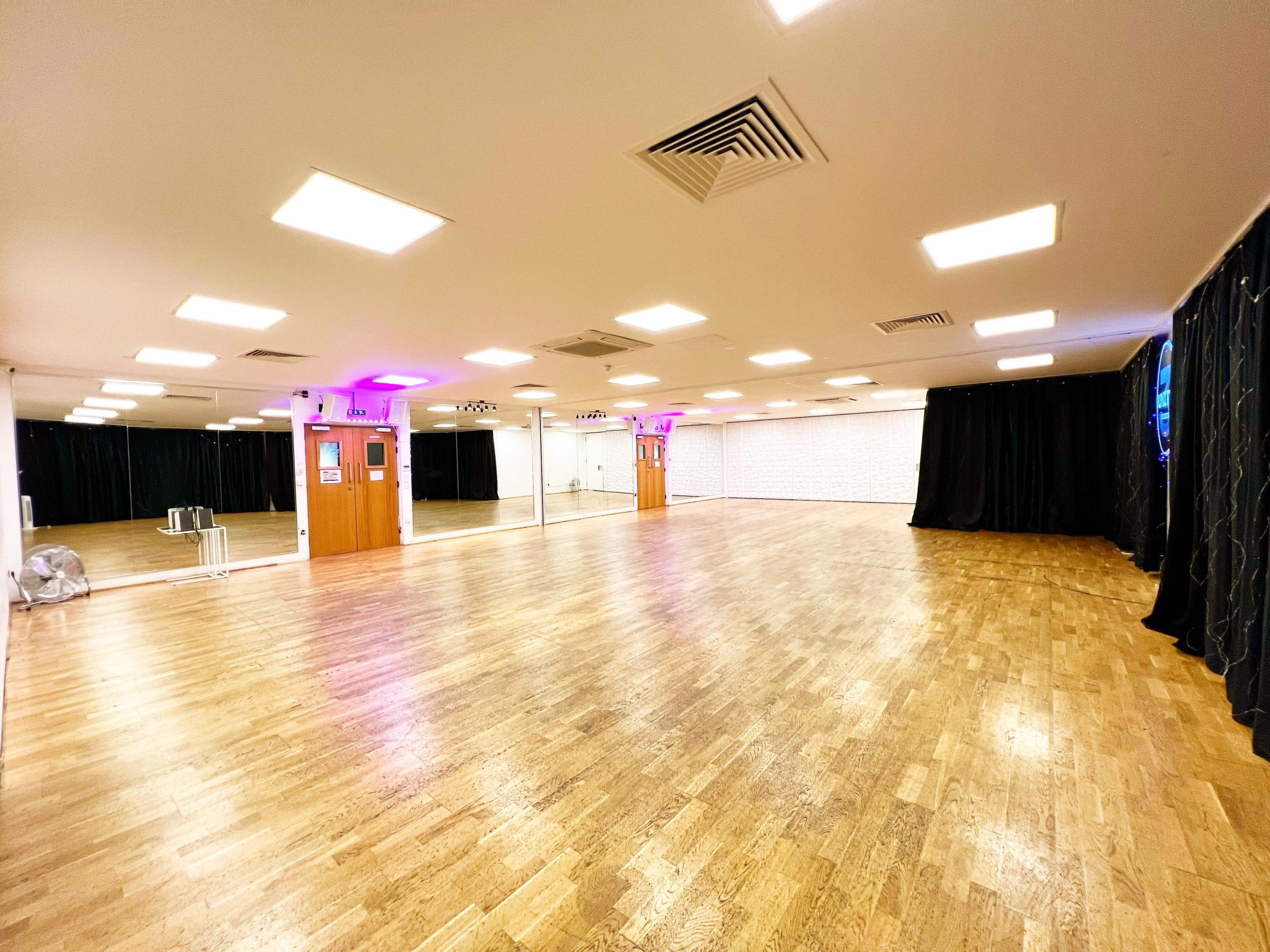 The image shows an empty dance studio with wooden flooring, wall mirrors, and soft lighting.