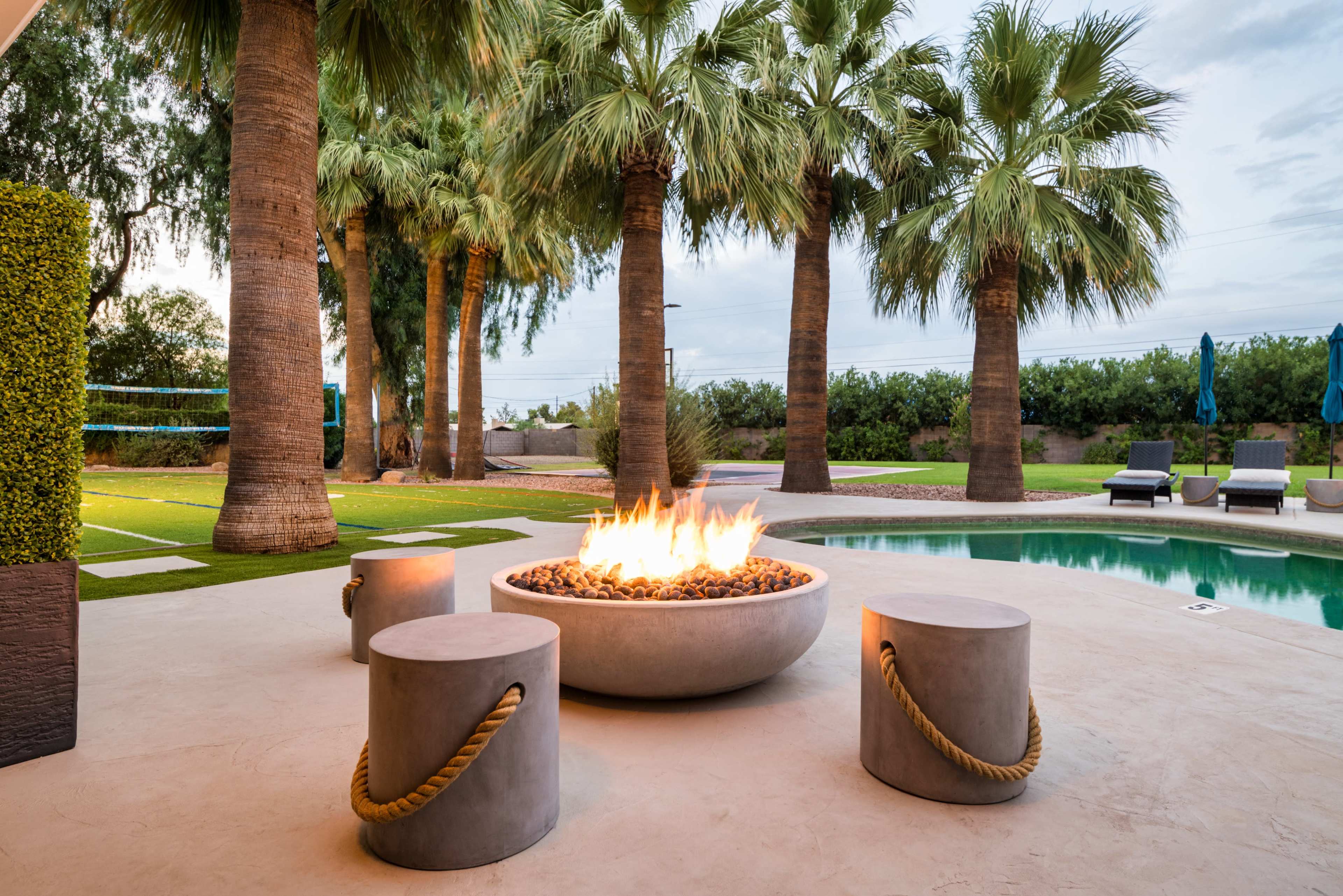 A modern outdoor space features a circular fire pit surrounded by cylindrical concrete stools, with palm trees and a swimming pool in the background.