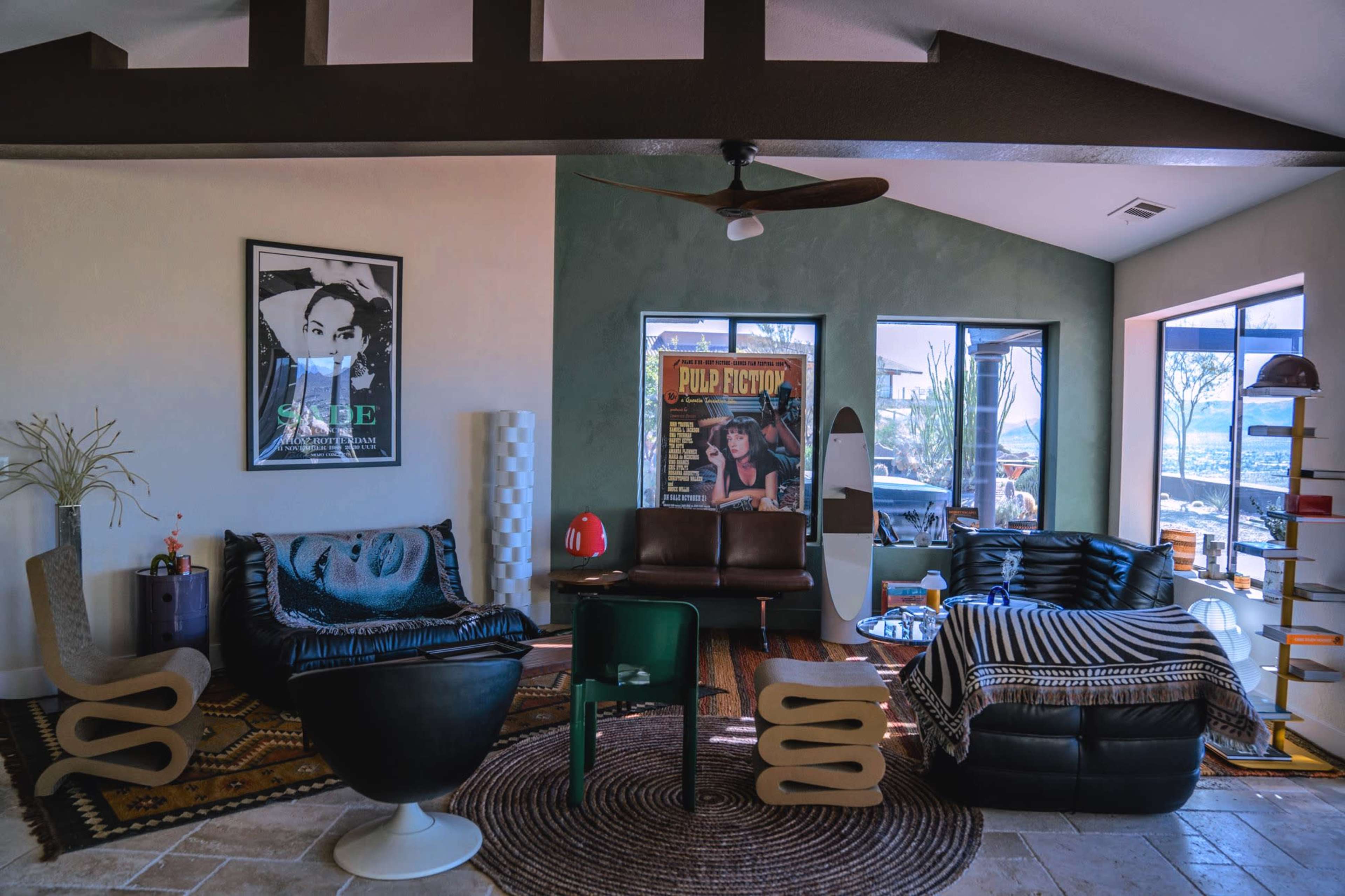 The living room features a mix of modern and vintage furniture, including a black leather sofa, a striped throw, and large windows that provide a view of the outdoors.