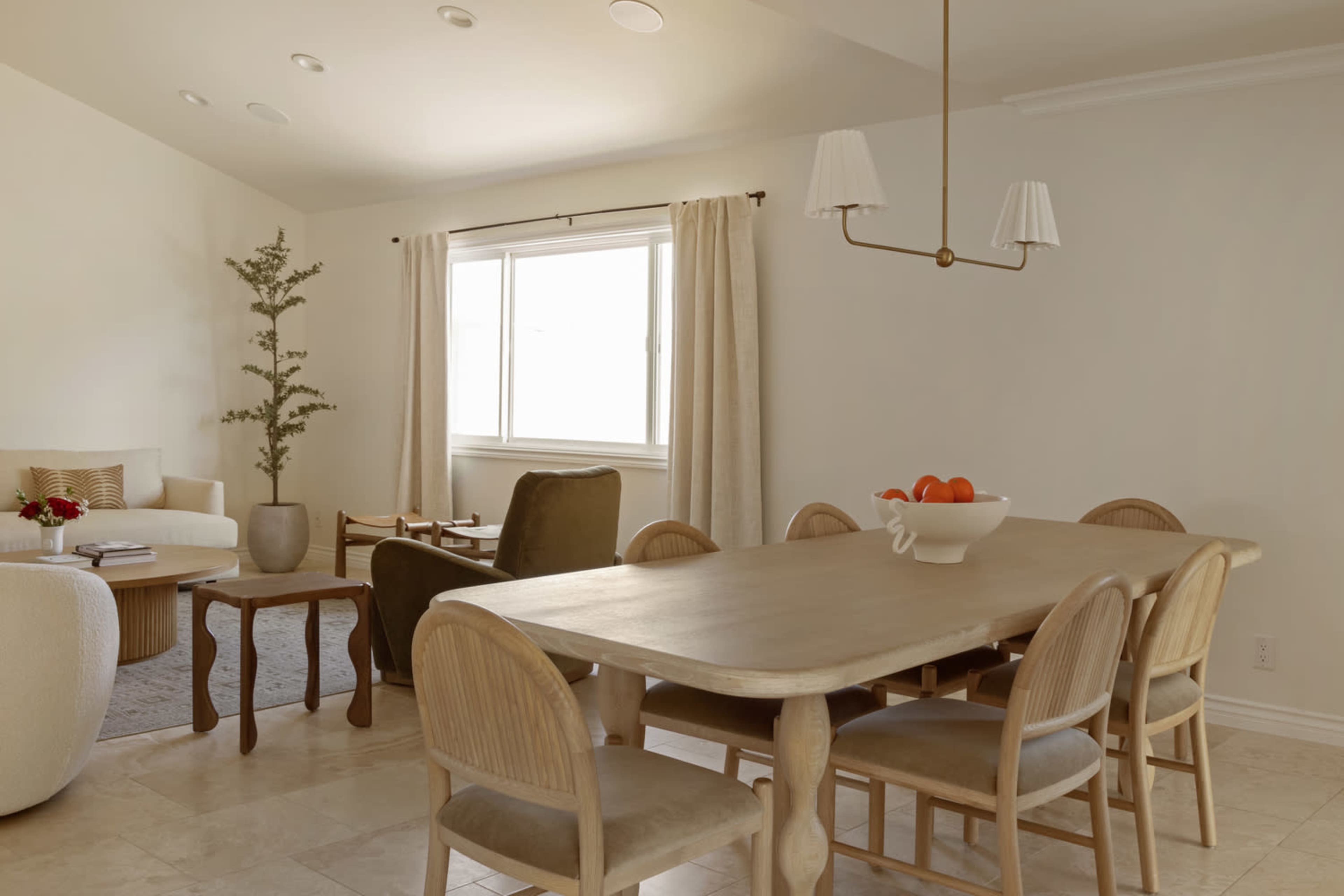 The image shows a dining area with a long wooden table surrounded by six curved-back chairs, a large window, and light decor elements, including a potted plant and a bowl of oranges on the table.