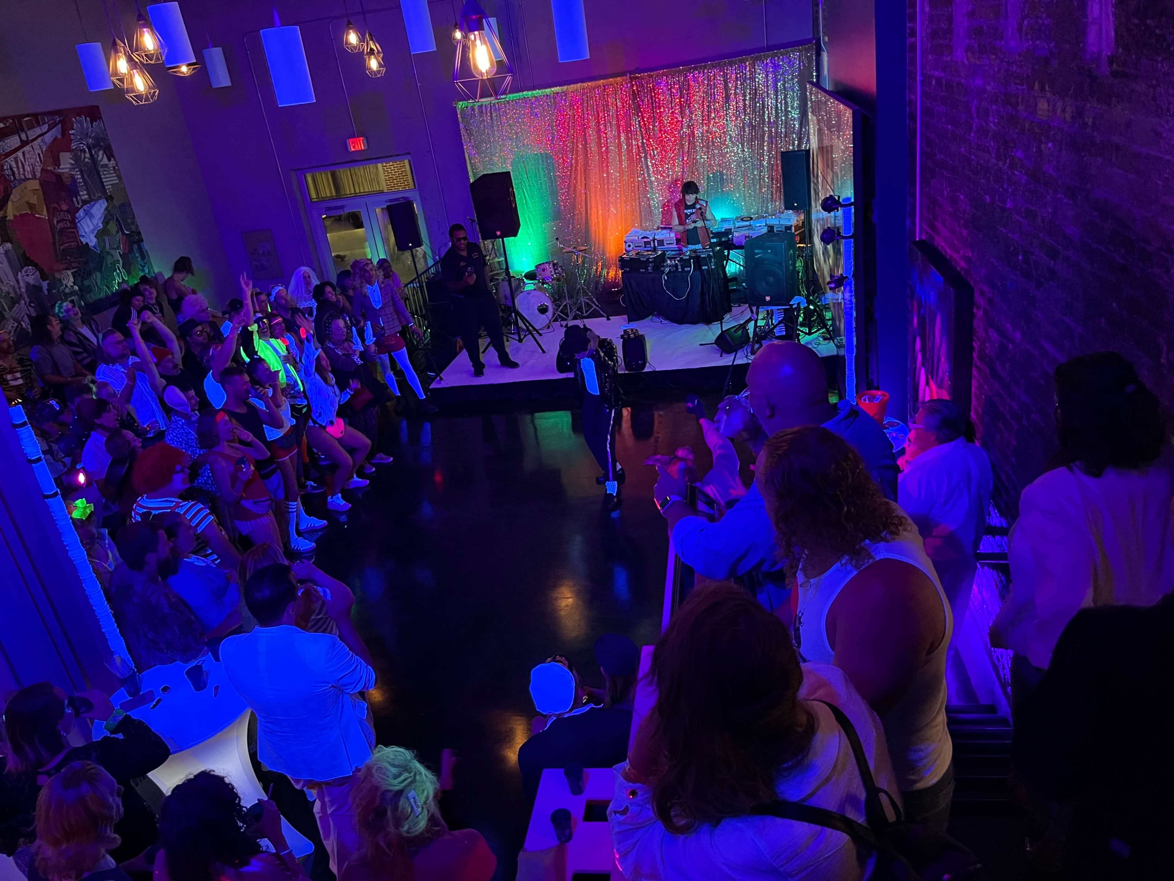 A large crowd gathers in a dimly lit venue, watching a performer on stage with colorful lights illuminating the space.