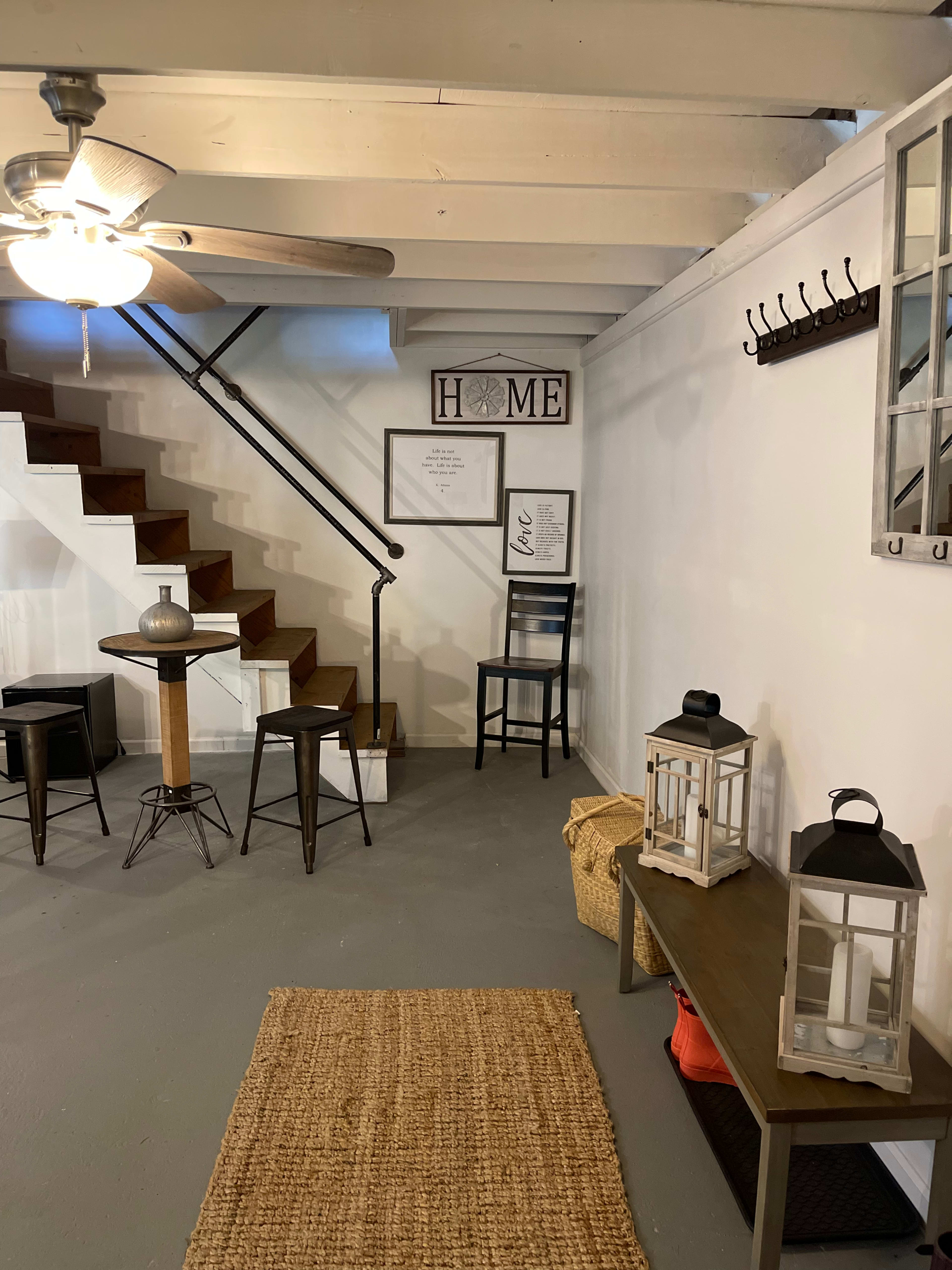 A basement area with a staircase, a small table and chairs, wall decor featuring the word "HOME," and a couple of lanterns on a console table.