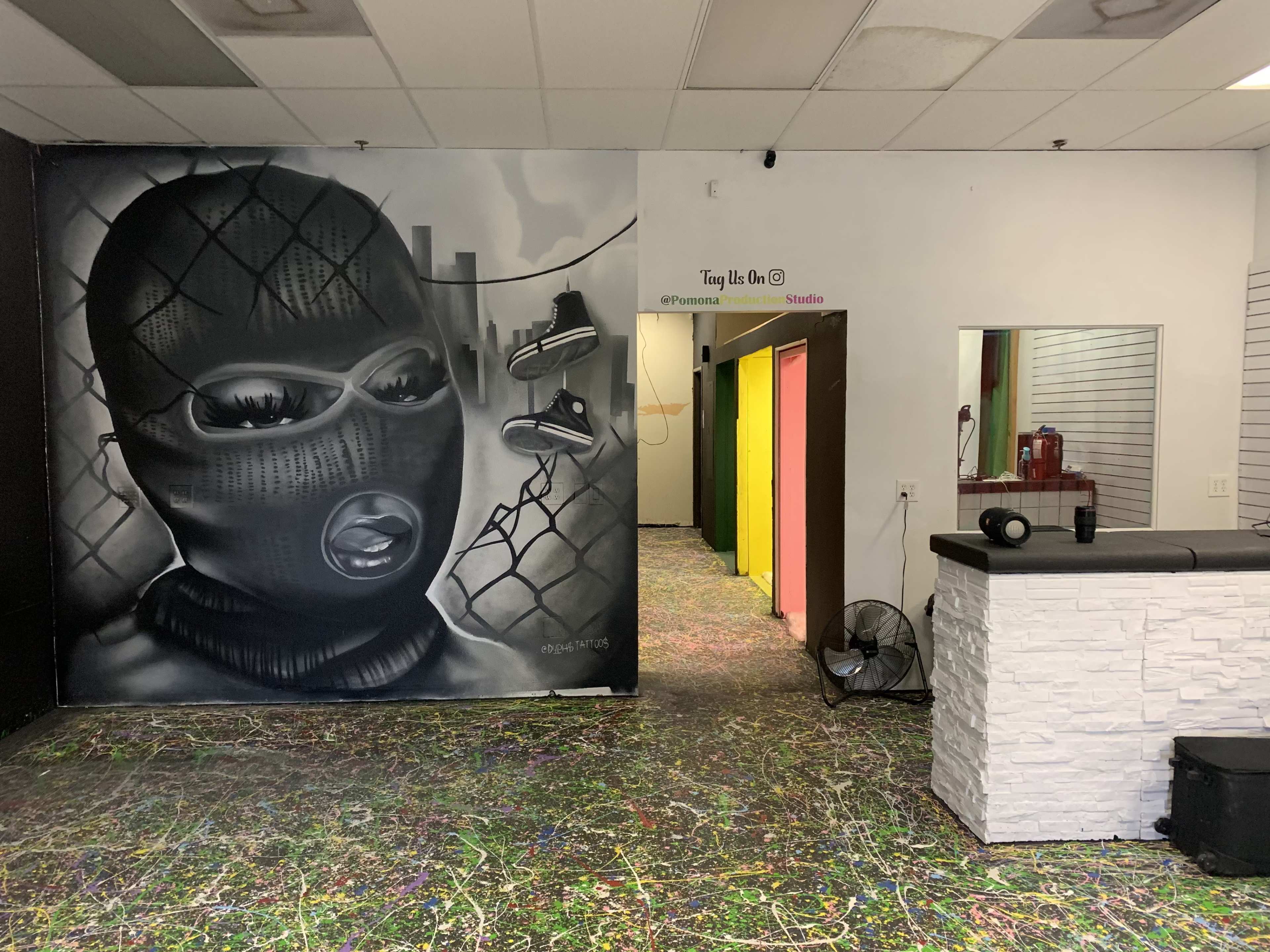 The image shows a colorful indoor space featuring a large mural of a person in a ski mask on one wall, with a hallway of doors painted in bright colors leading to another area.