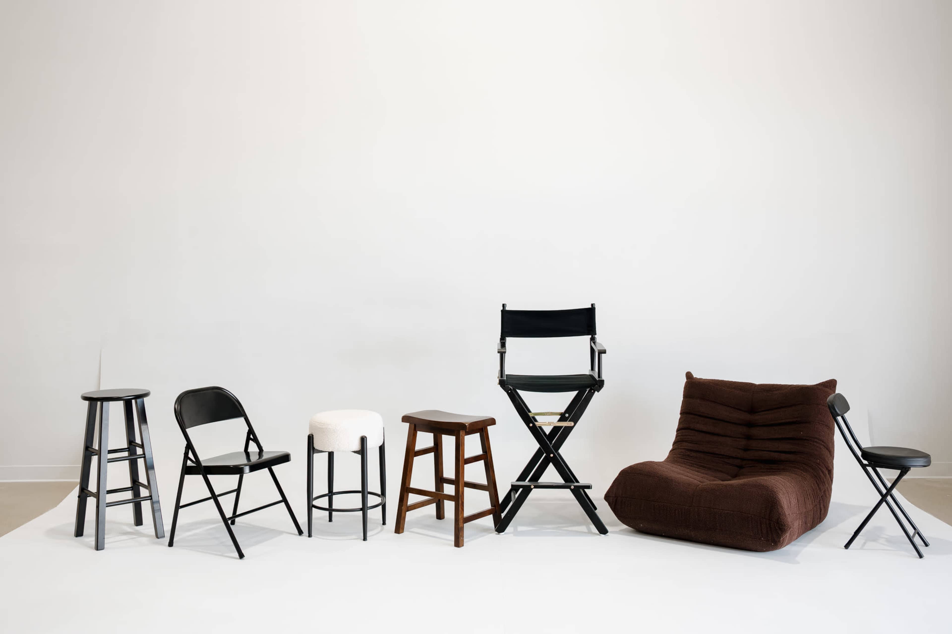 A variety of chairs and stools in different styles and materials are arranged against a plain white wall.
