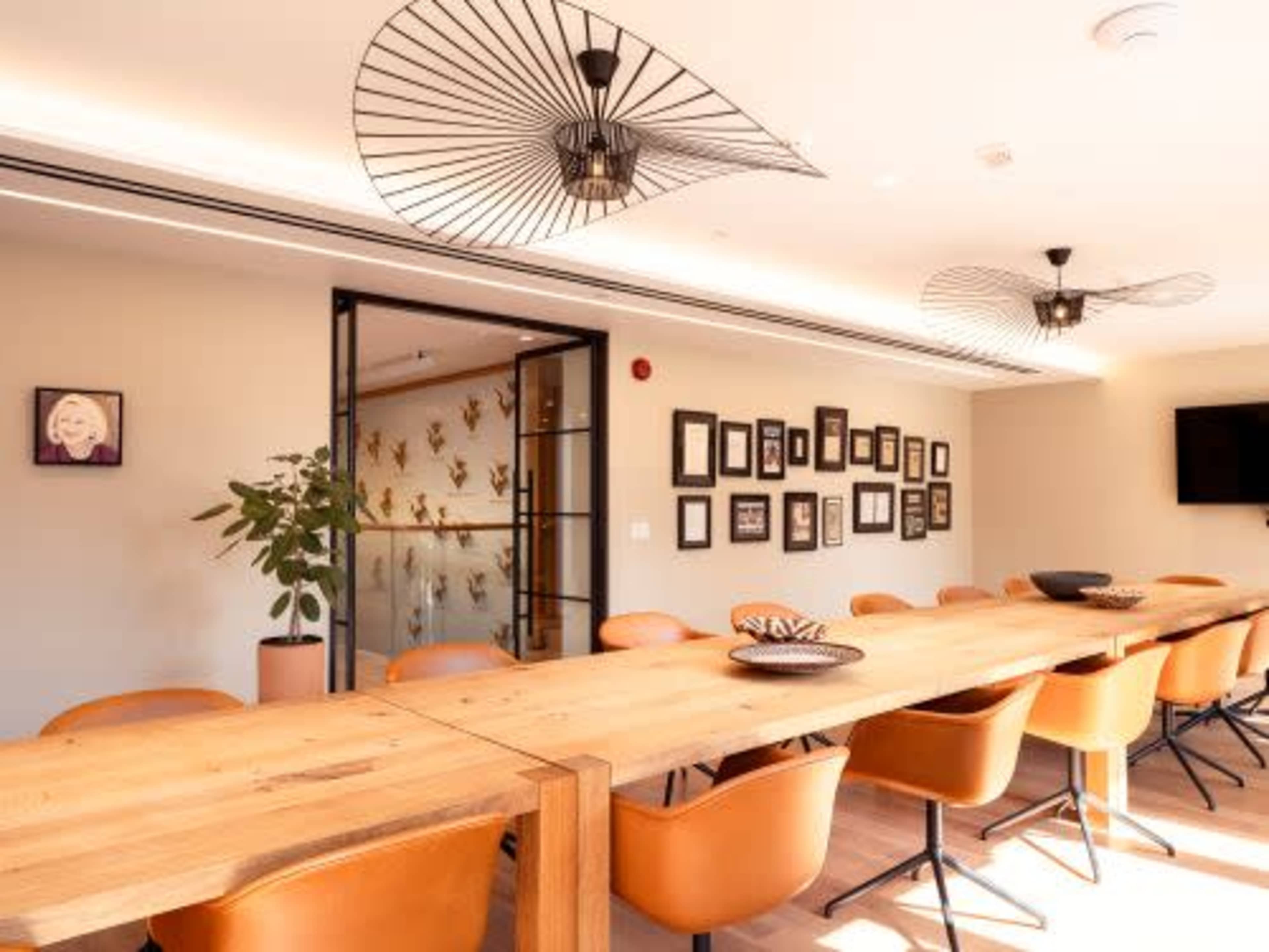 The image shows a modern dining area with a long wooden table surrounded by orange chairs and framed photographs on the wall.