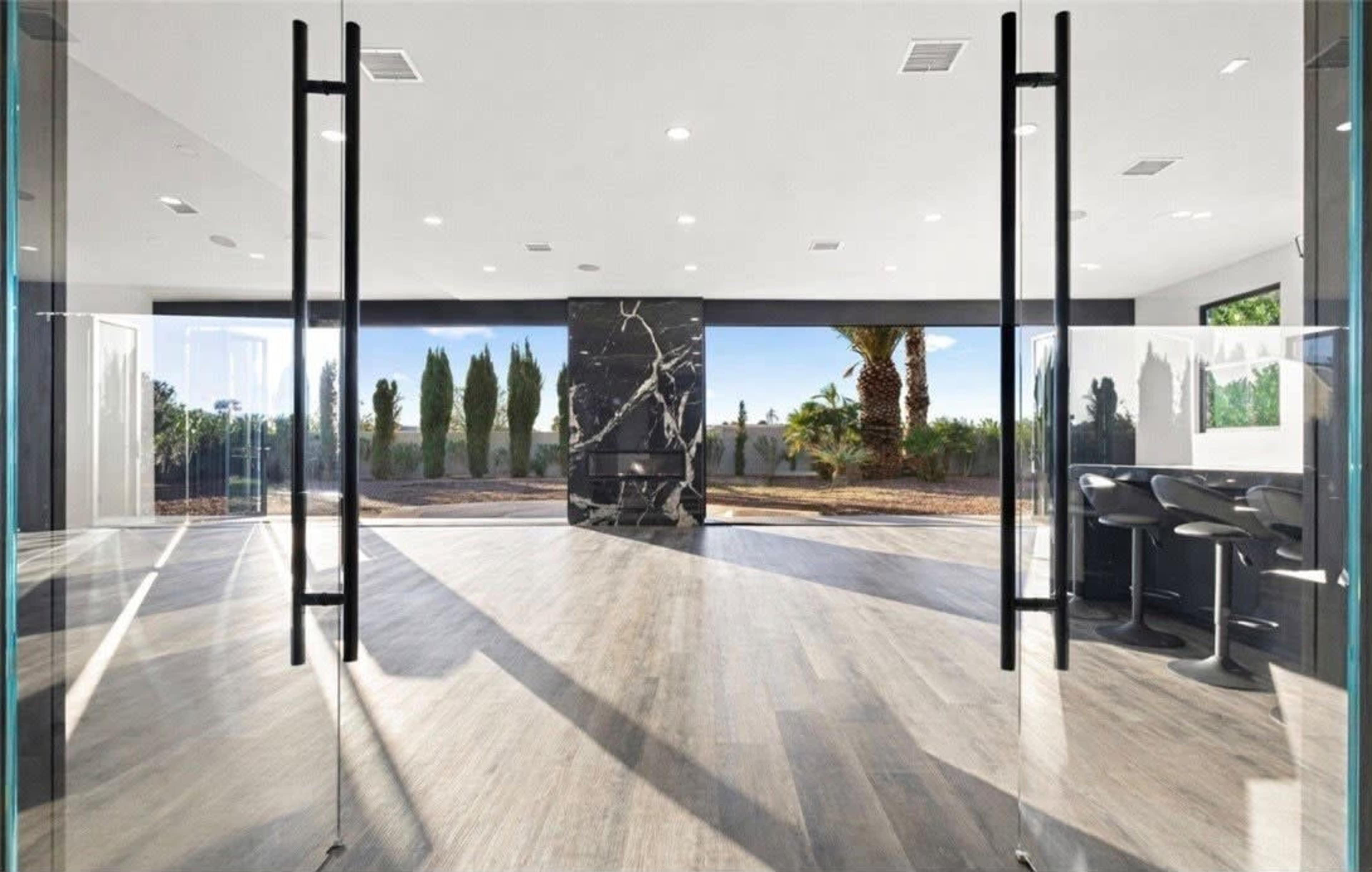 The image shows a modern interior space with large glass doors opening to a landscaped view featuring palm trees and a dark marble feature wall.