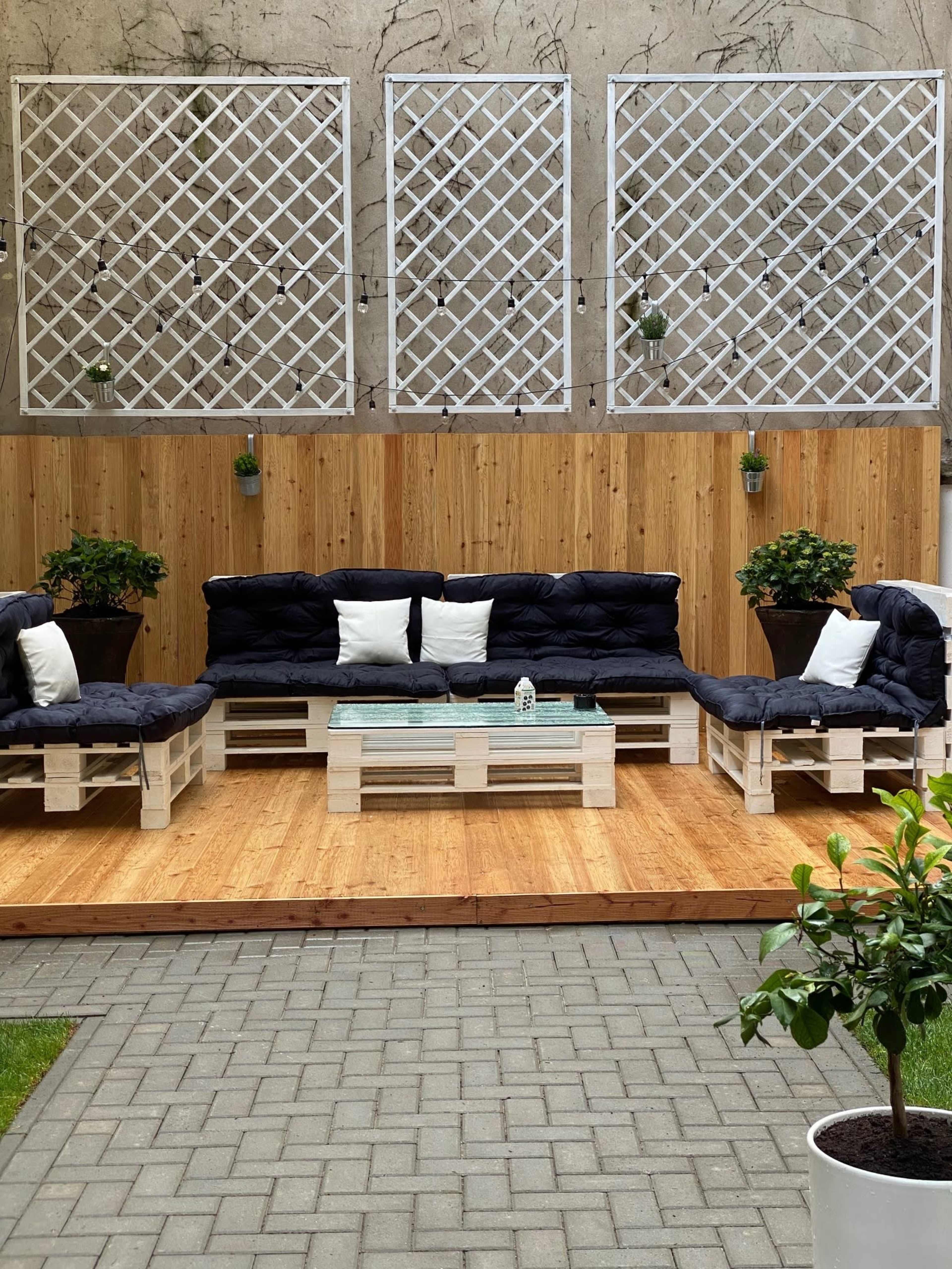 A cozy outdoor seating area with wooden pallet furniture, featuring blue cushions, a glass-top coffee table, and potted plants against a wooden backdrop.