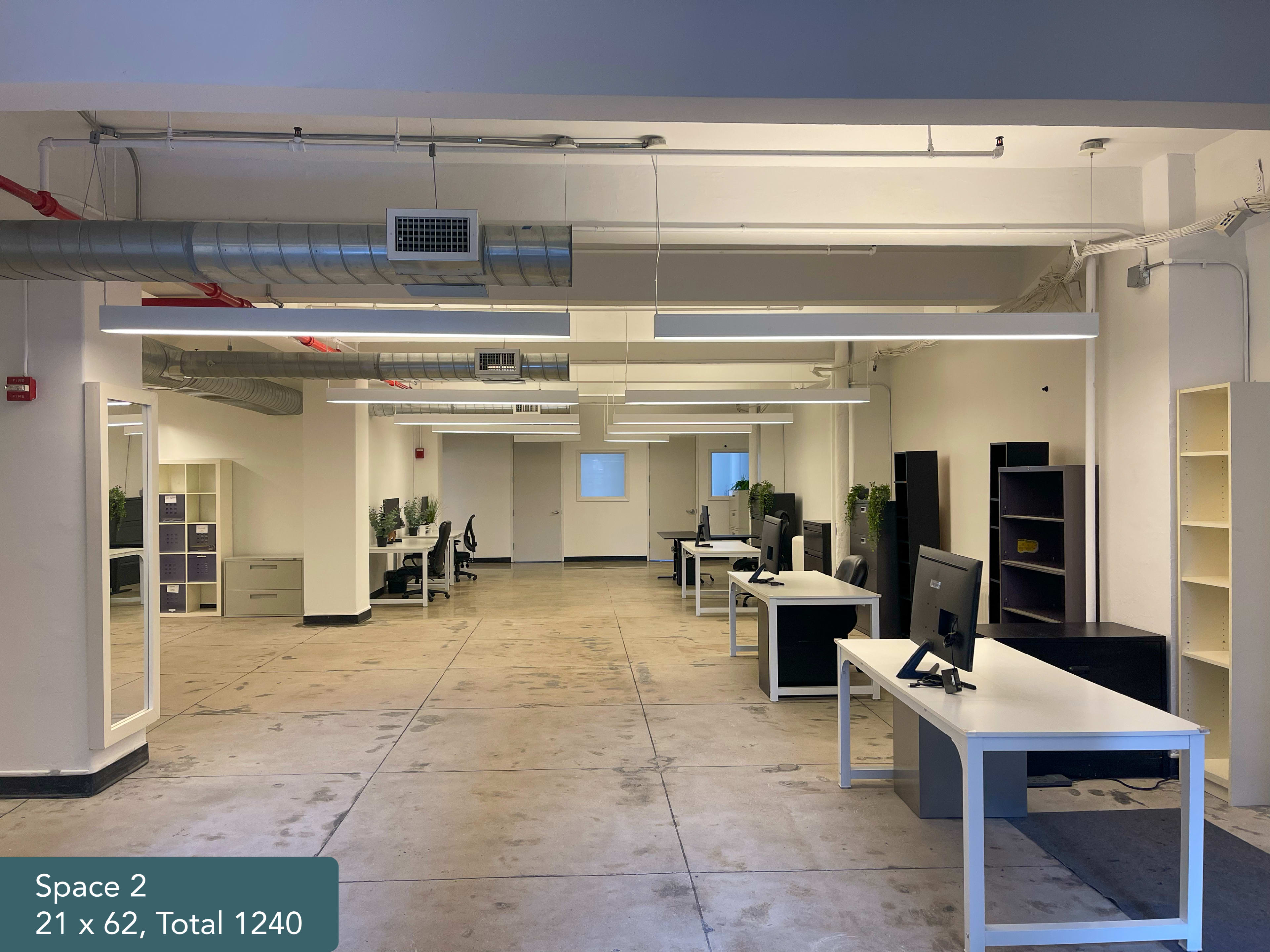 The image shows a spacious office area with multiple white desks equipped with computers and plants, illuminated by overhead LED lights.