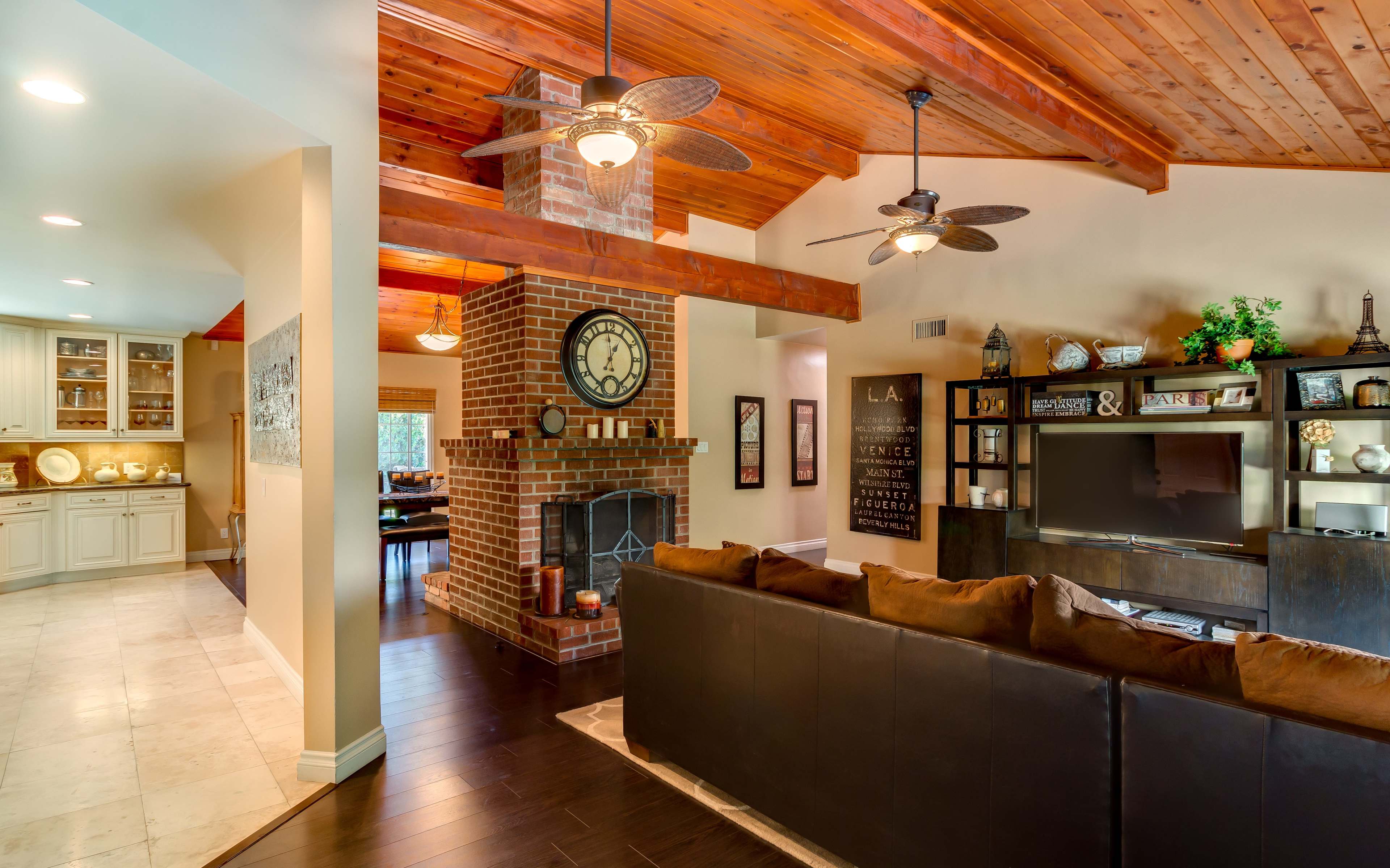 A modern living space featuring a brown sectional sofa, a brick fireplace with a clock, and wooden beams overhead, seamlessly connecting to a dining area with a kitchen.