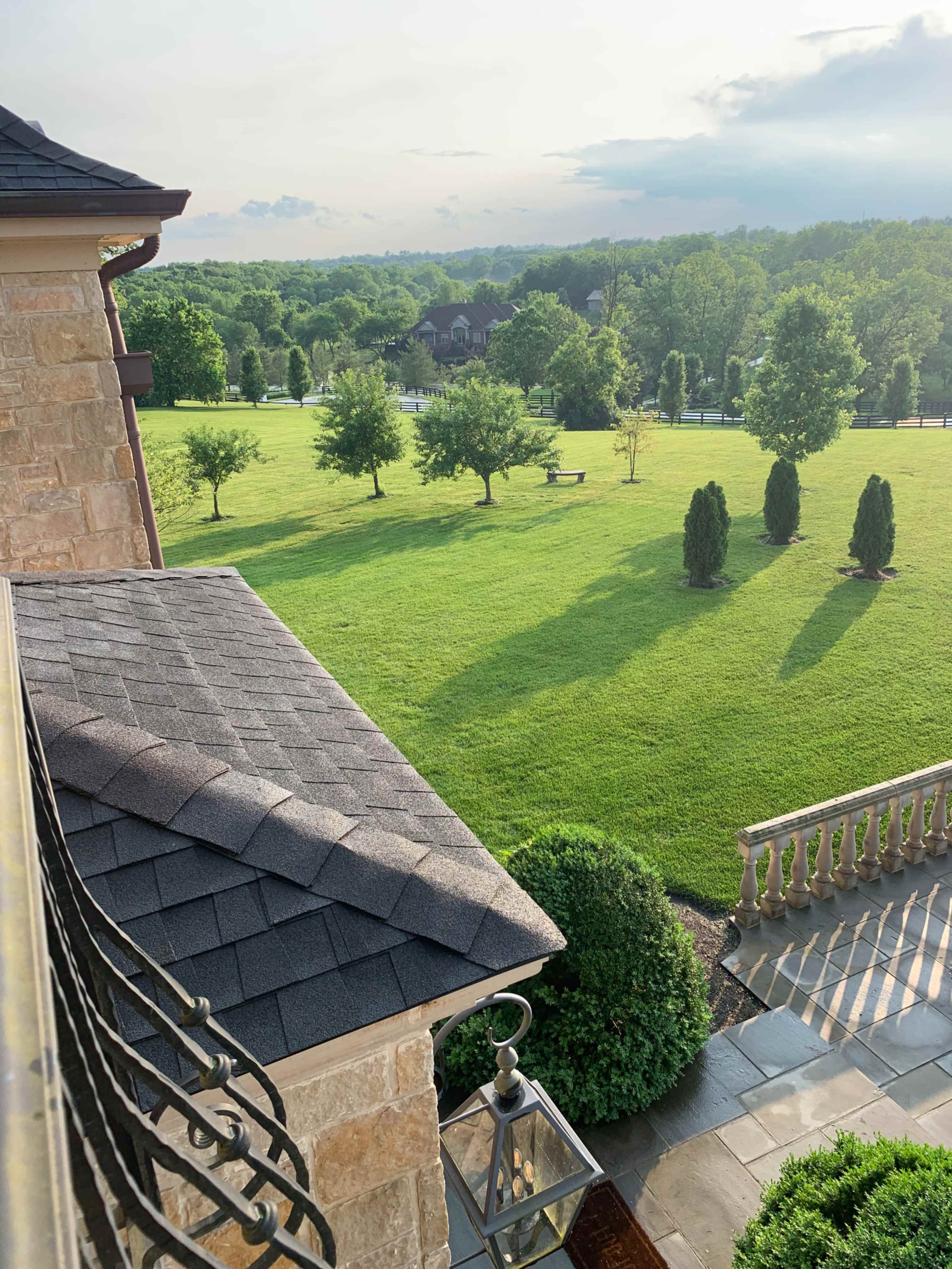 A view from a balcony overlooking a lush green lawn with several trees and a large field in the background.