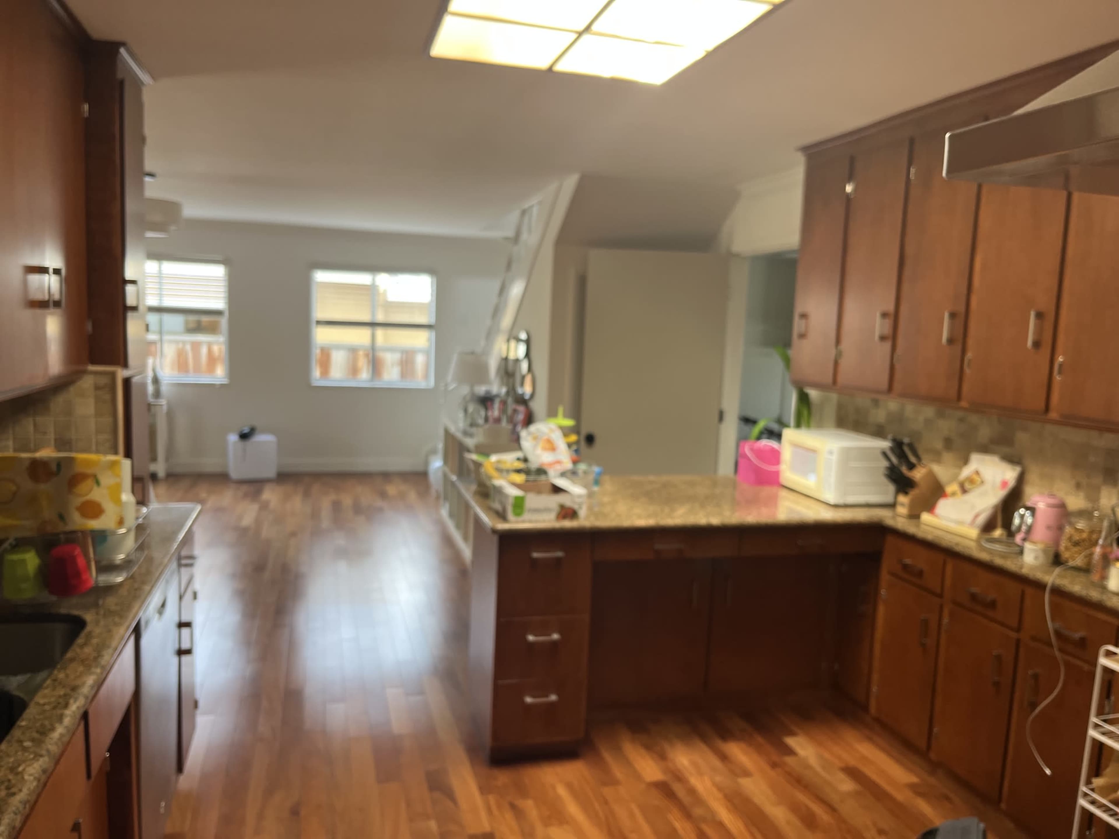 The image shows a spacious kitchen with wooden cabinets and granite countertops, leading into a well-lit adjoining room.