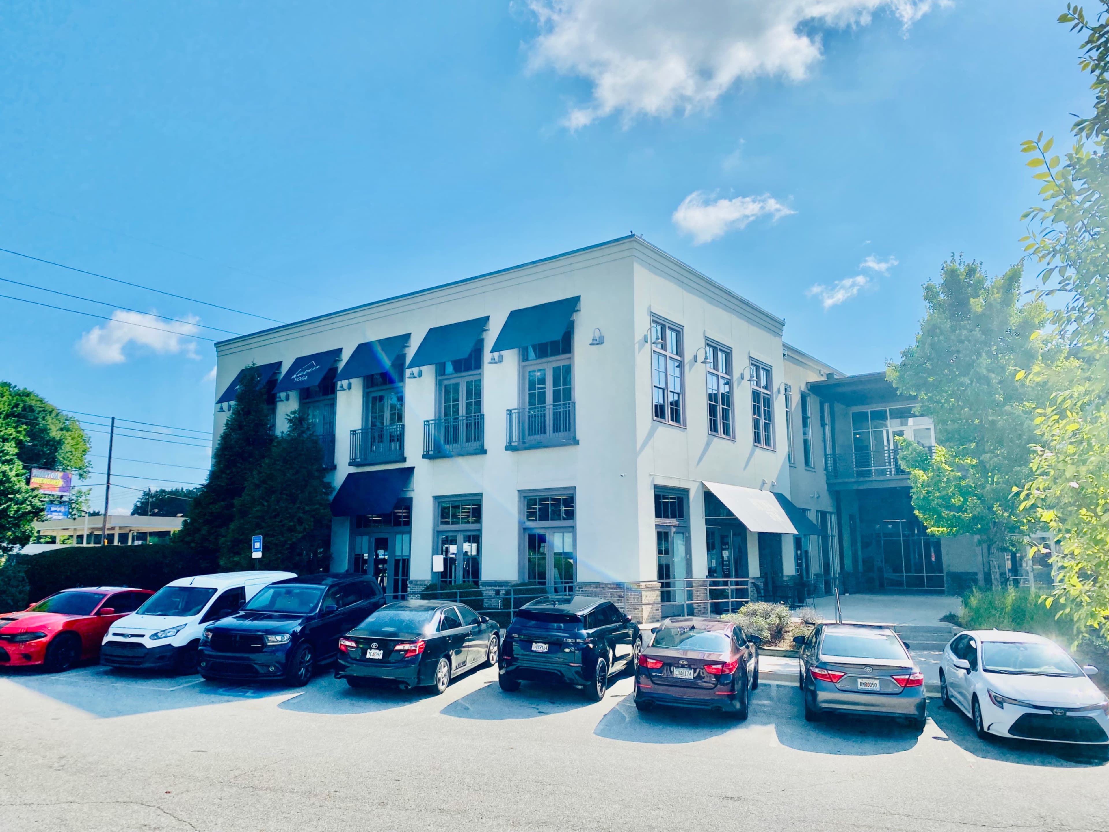 A modern commercial building is flanked by several parked cars in a parking lot under a clear blue sky.