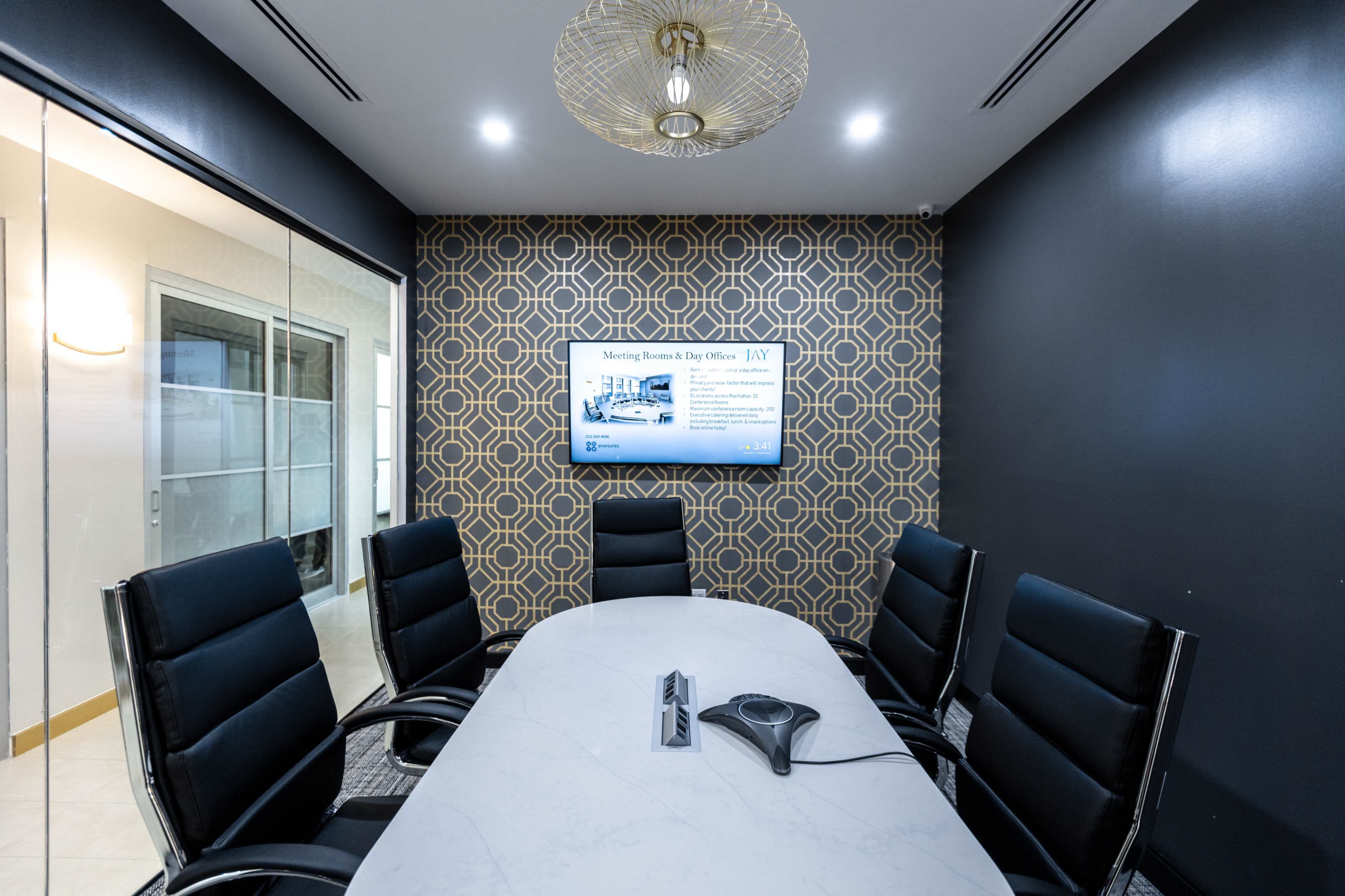 A modern conference room with a round table, six black leather chairs, and a wall-mounted screen displaying presentation content.