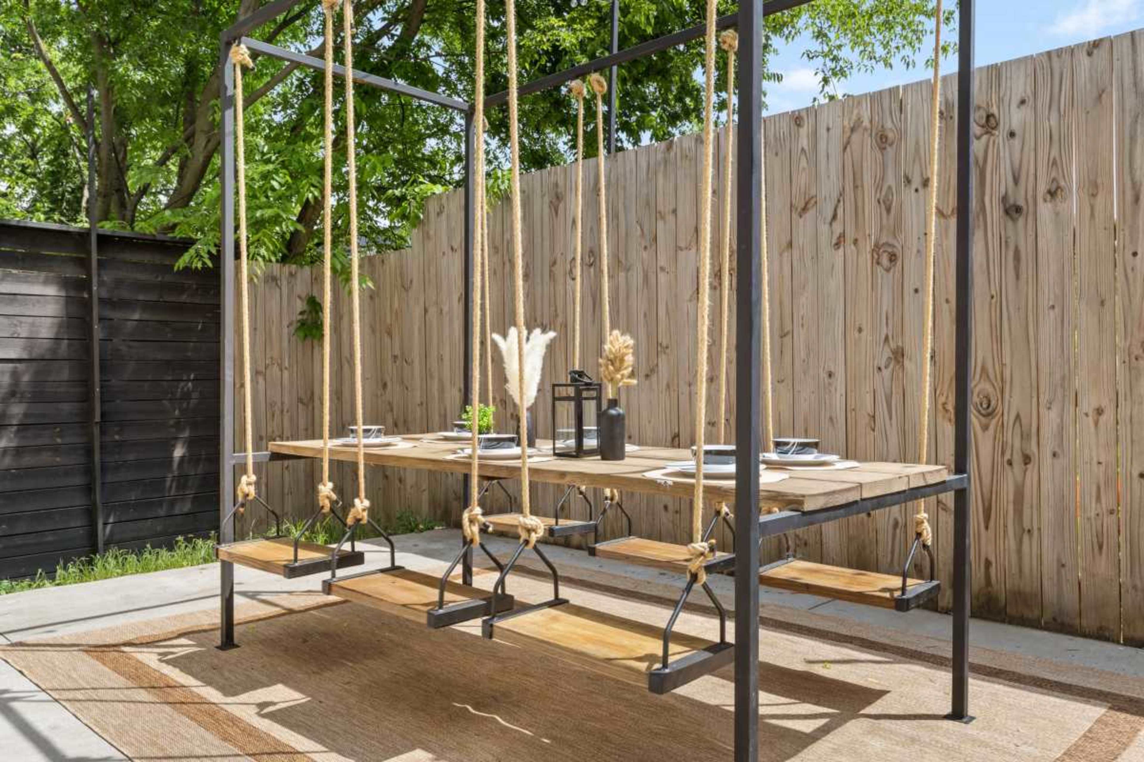 The image shows a modern outdoor dining table suspended from a frame with wooden swings as seats, set against a wooden fence and greenery.