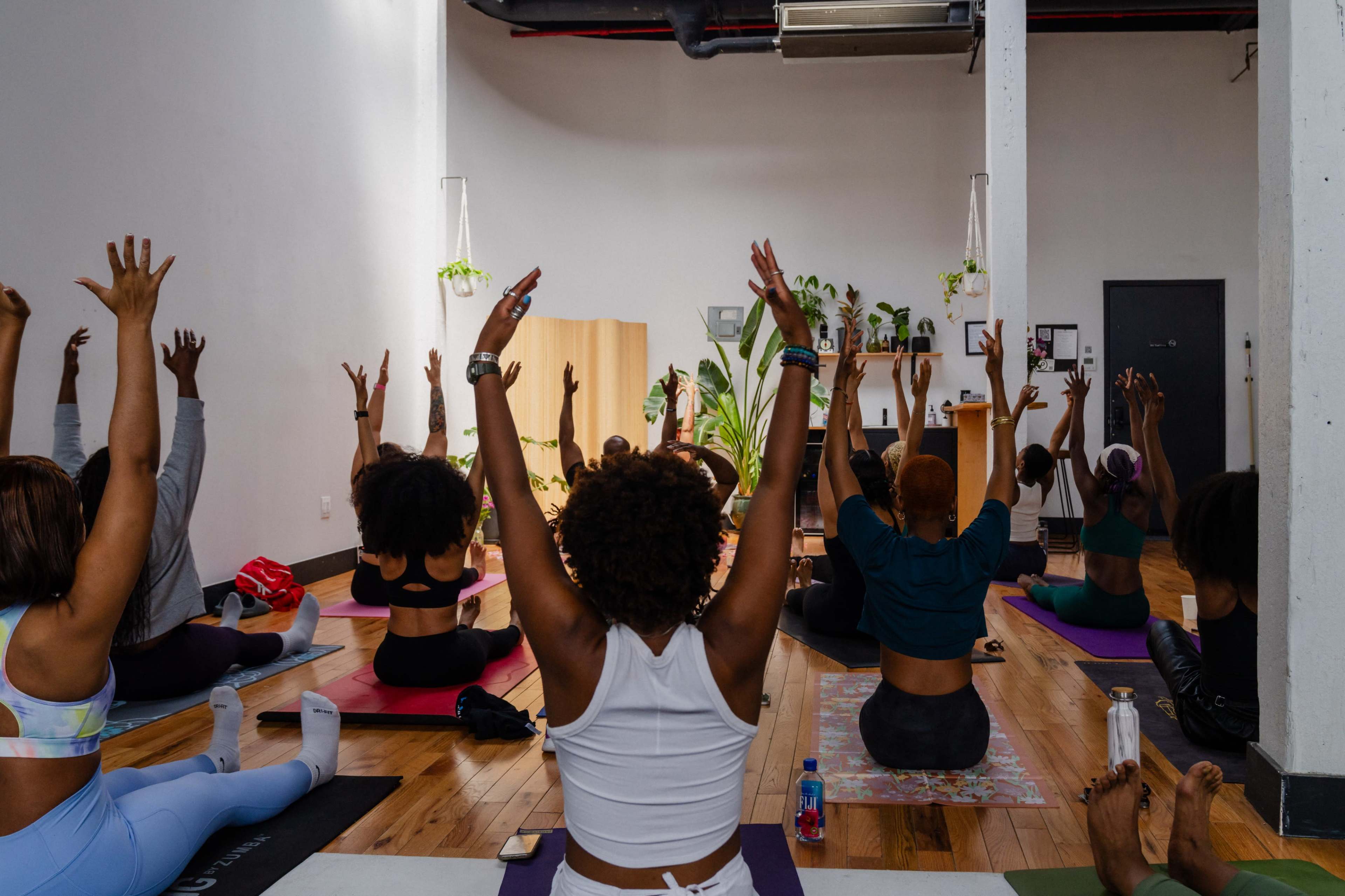 A group of people practice yoga in a spacious studio, reaching their arms upward while seated on colorful mats.