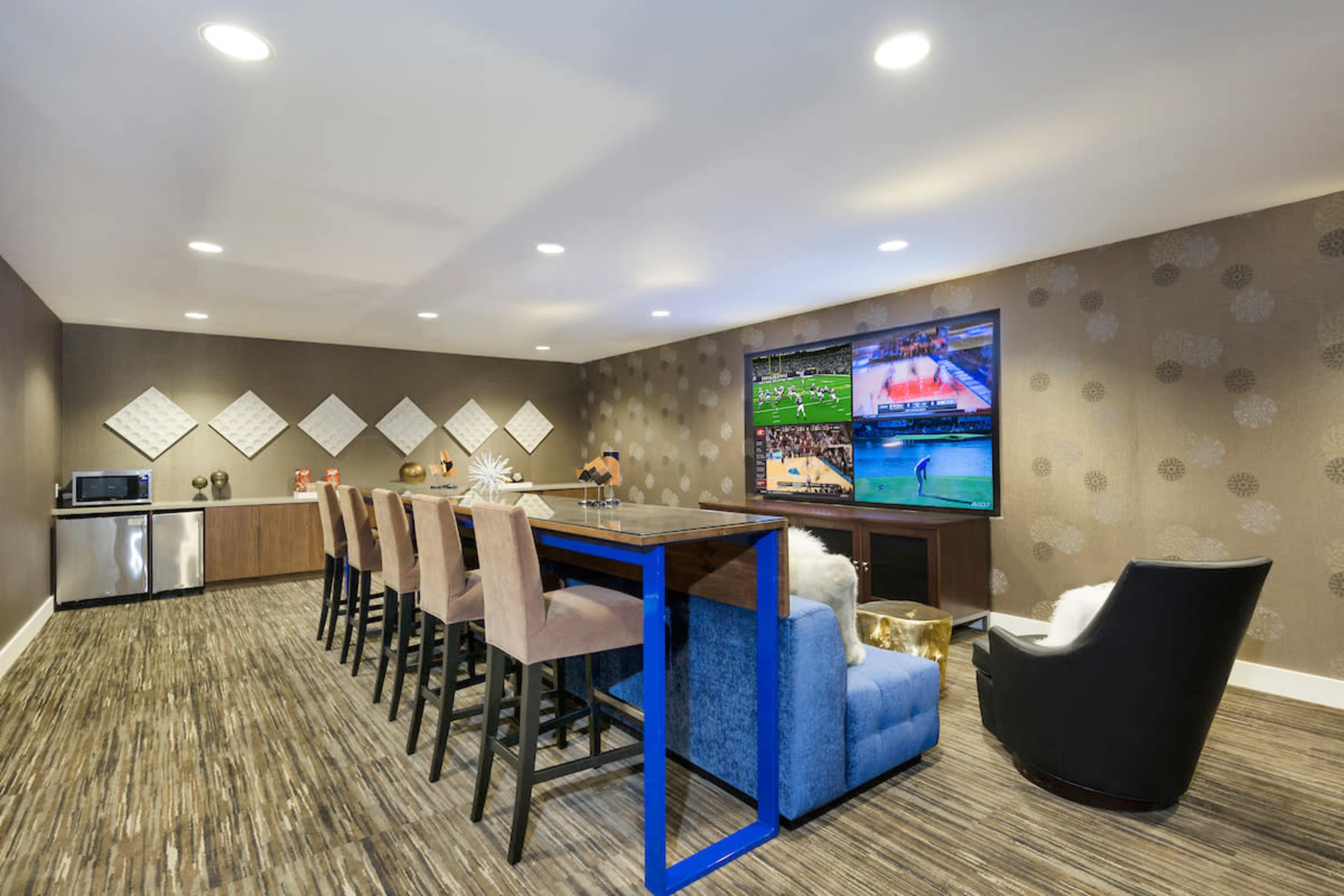 The image shows a modern entertainment room with a large television displaying multiple sports games, a bar-style table with stools, and a cozy seating area.