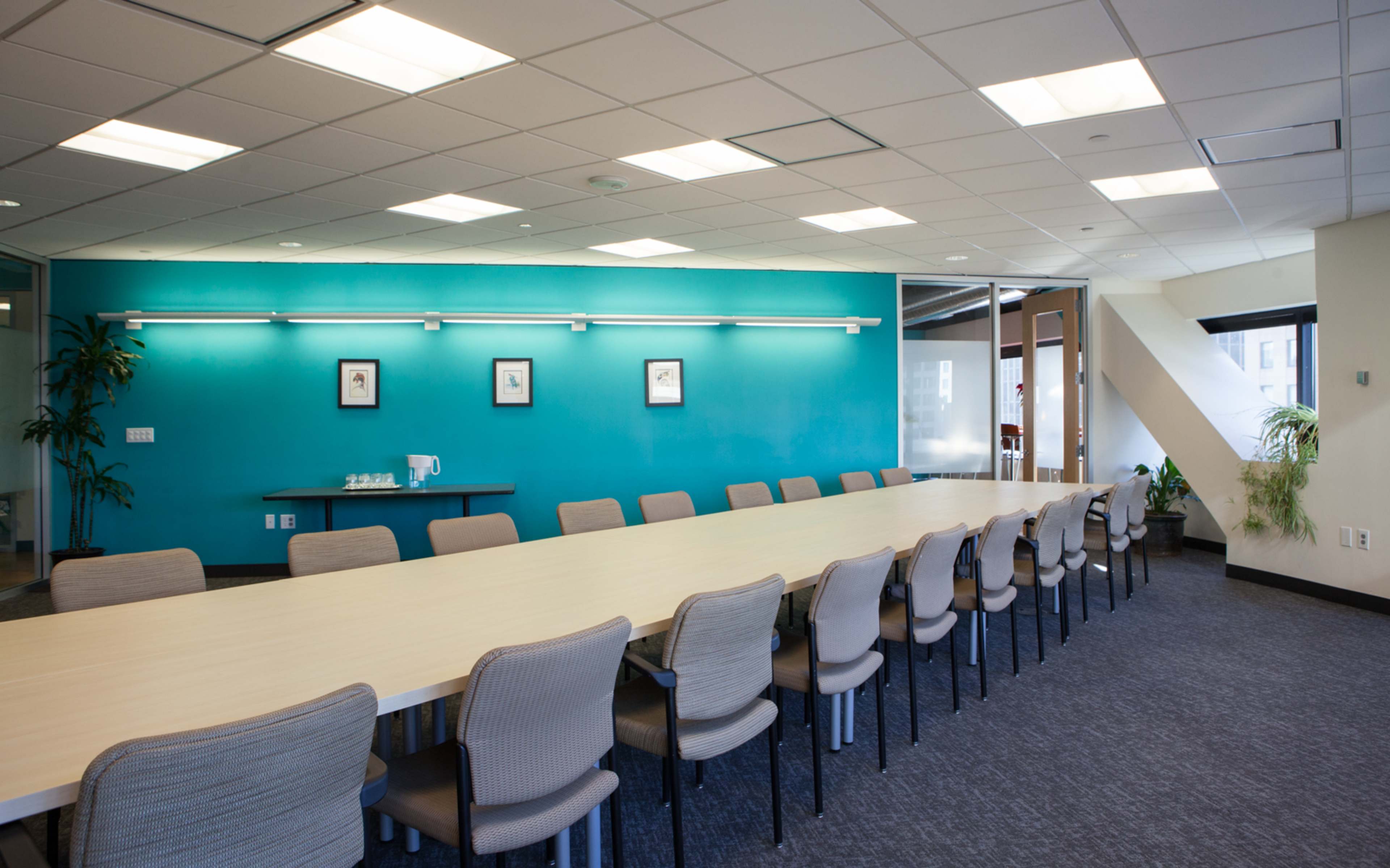 A long conference table is set in a well-lit meeting room with a turquoise accent wall and several chairs arranged along one side.