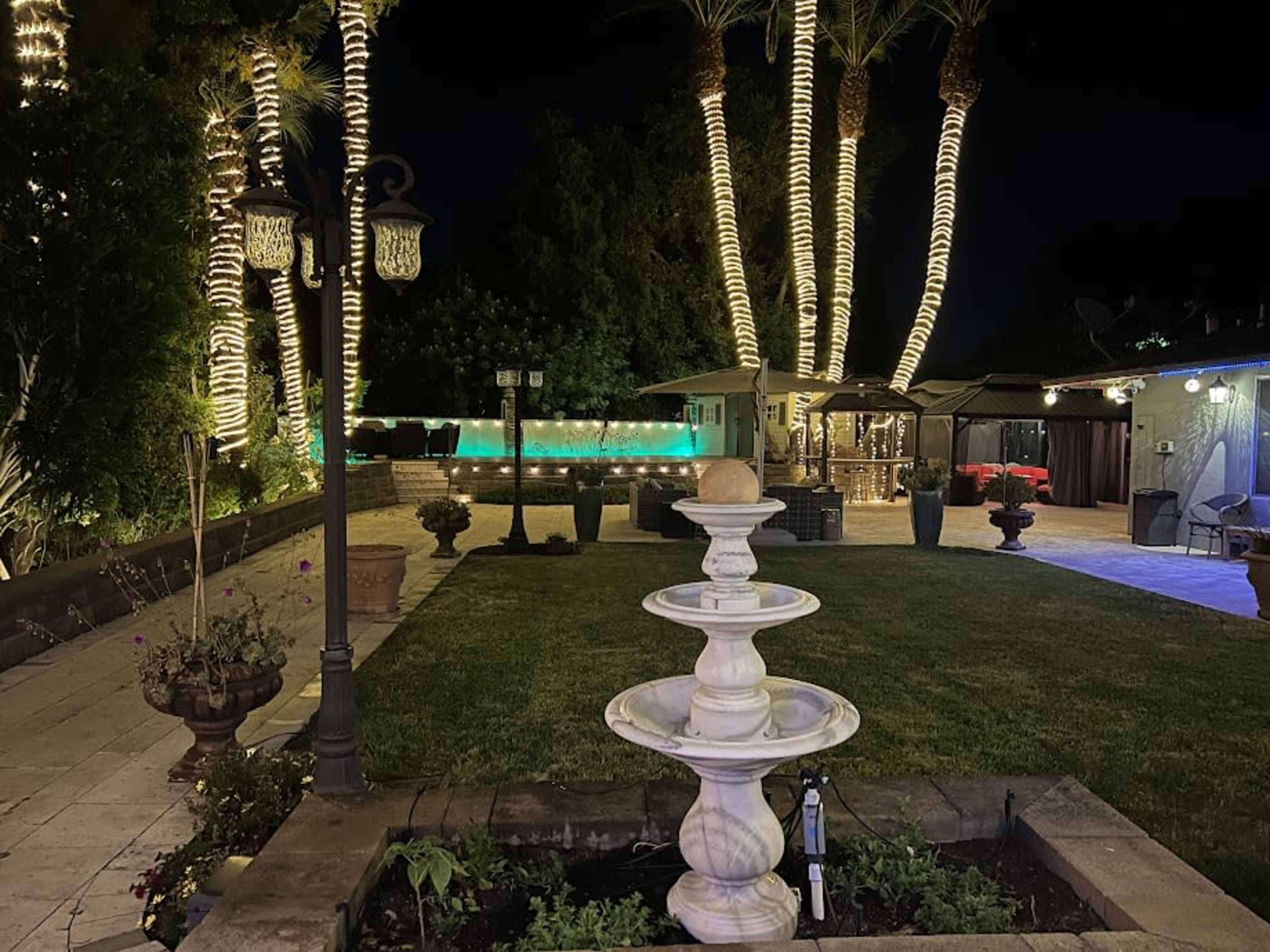 The image shows a backyard scene at night featuring a stone fountain in the foreground, palm trees wrapped in lights, and a pool area illuminated by soft lighting.