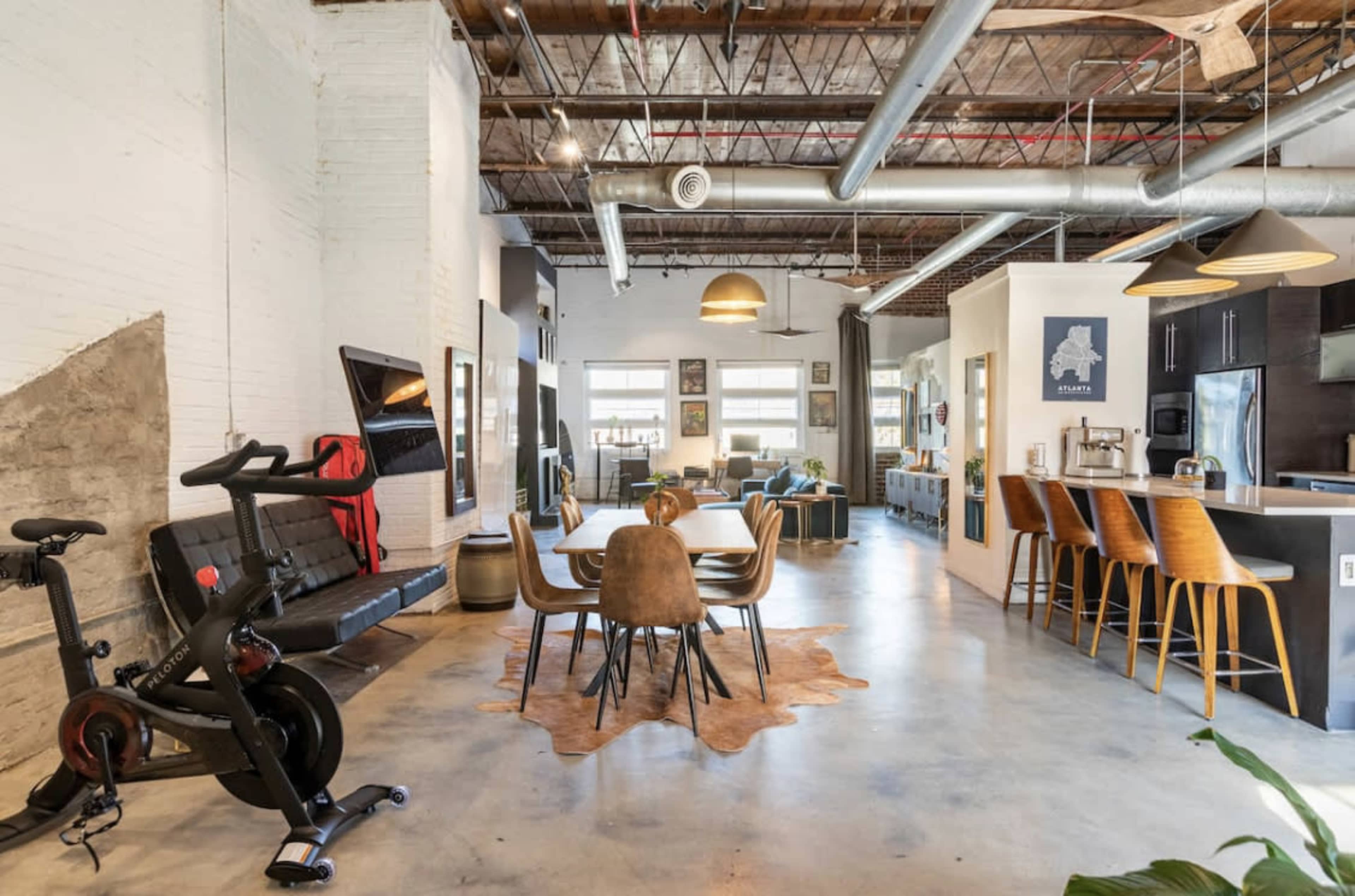 A spacious, modern open-concept interior featuring a dining area with a wooden table, chairs, and a bike, alongside a kitchen and lounge area.