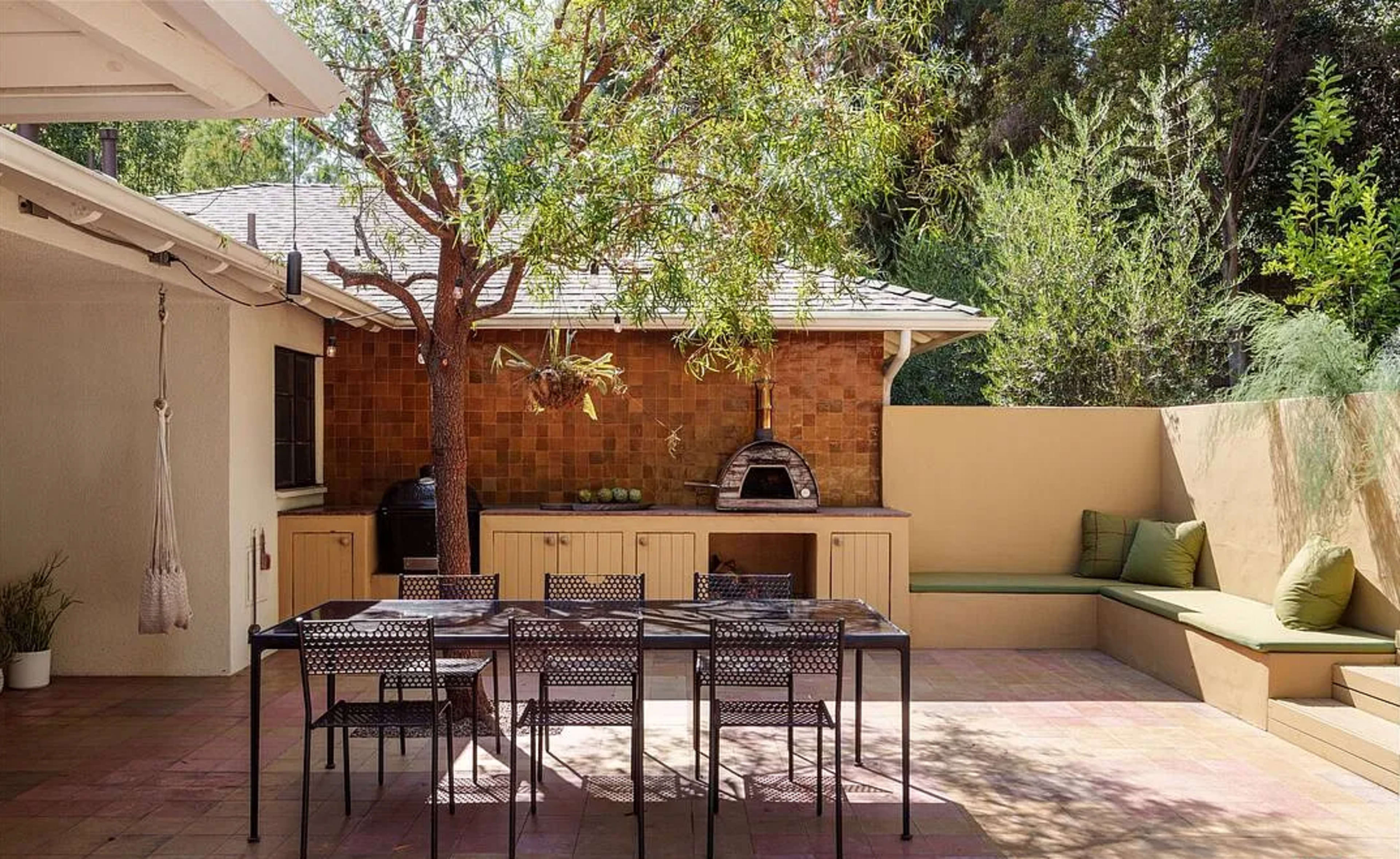 An outdoor dining area features a large table, an outdoor kitchen with a pizza oven, and seating surrounded by greenery.