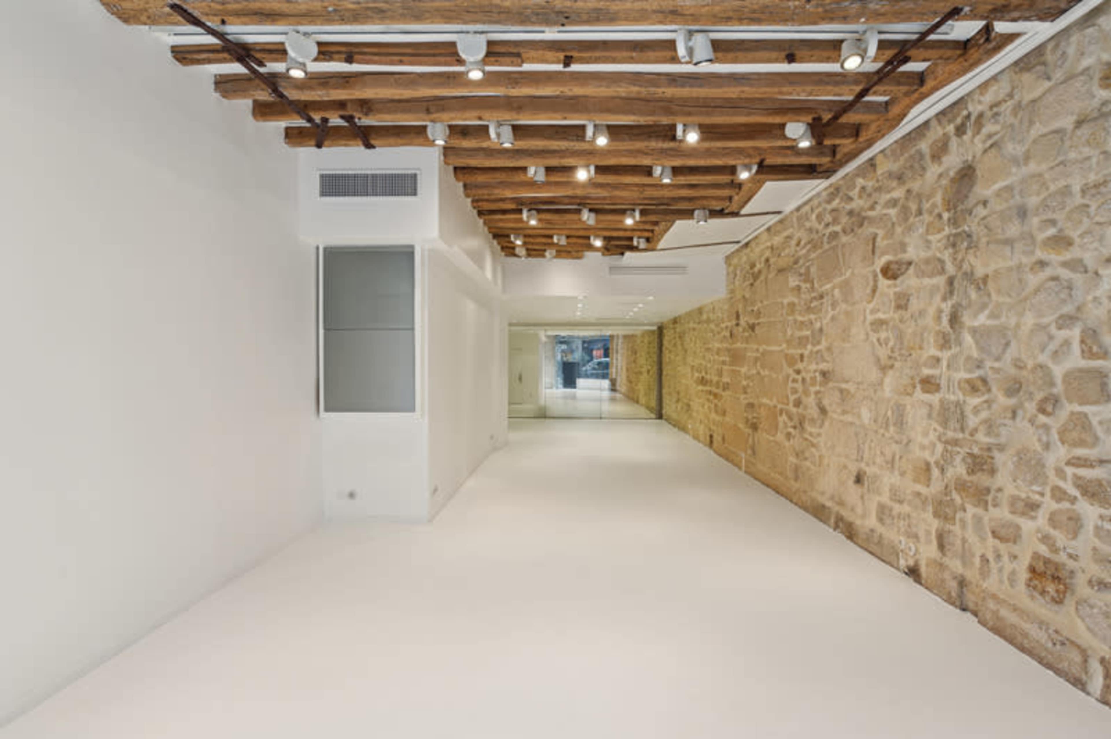 The image shows a minimalist interior space with a stone wall, wooden beams on the ceiling, and white flooring.
